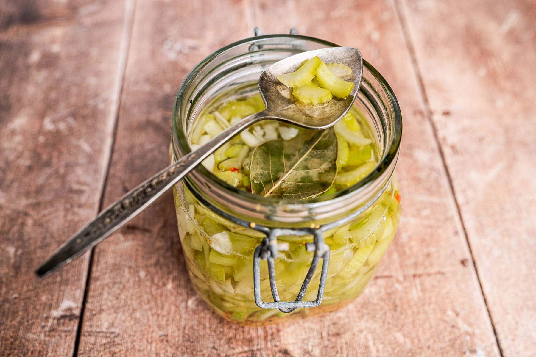 A glass jar filled with pickled celery and a bay leaf sits on a wooden surface, with a metal spoon resting on top, holding tangy pickled celery slices.