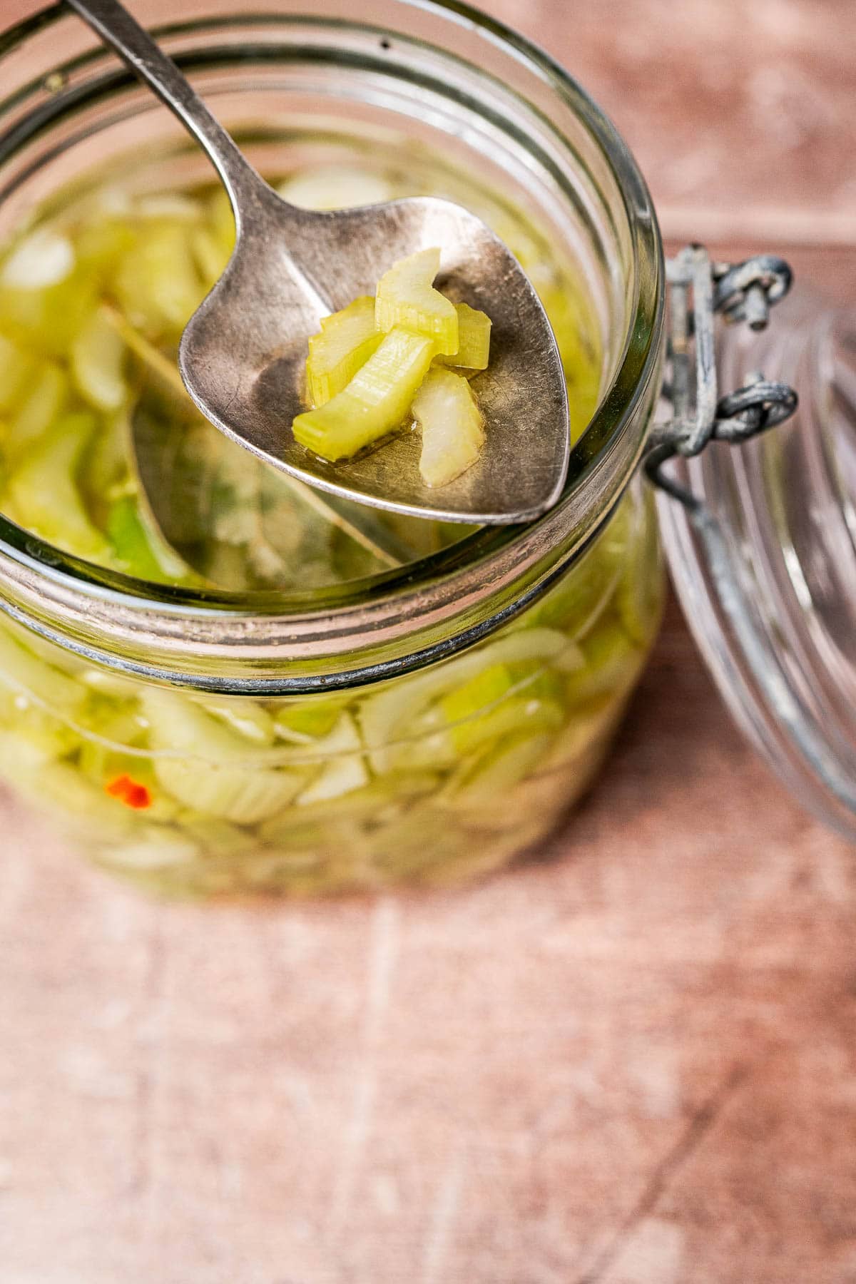 A spoon holding chopped pickled celery above an open glass jar filled with tangy pickled celery pieces.