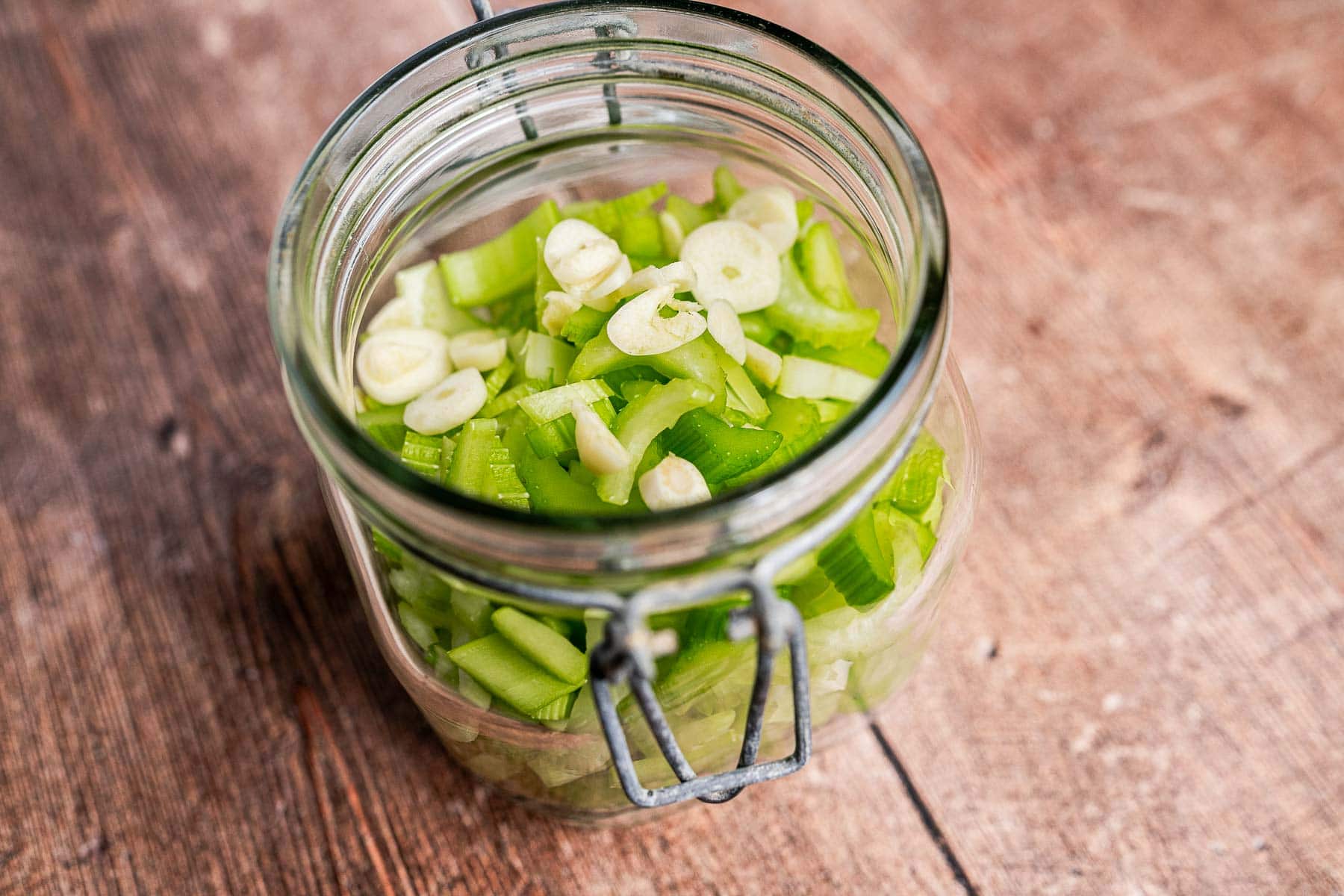 A glass jar filled with pickled celery and sliced garlic sits on a wooden surface.