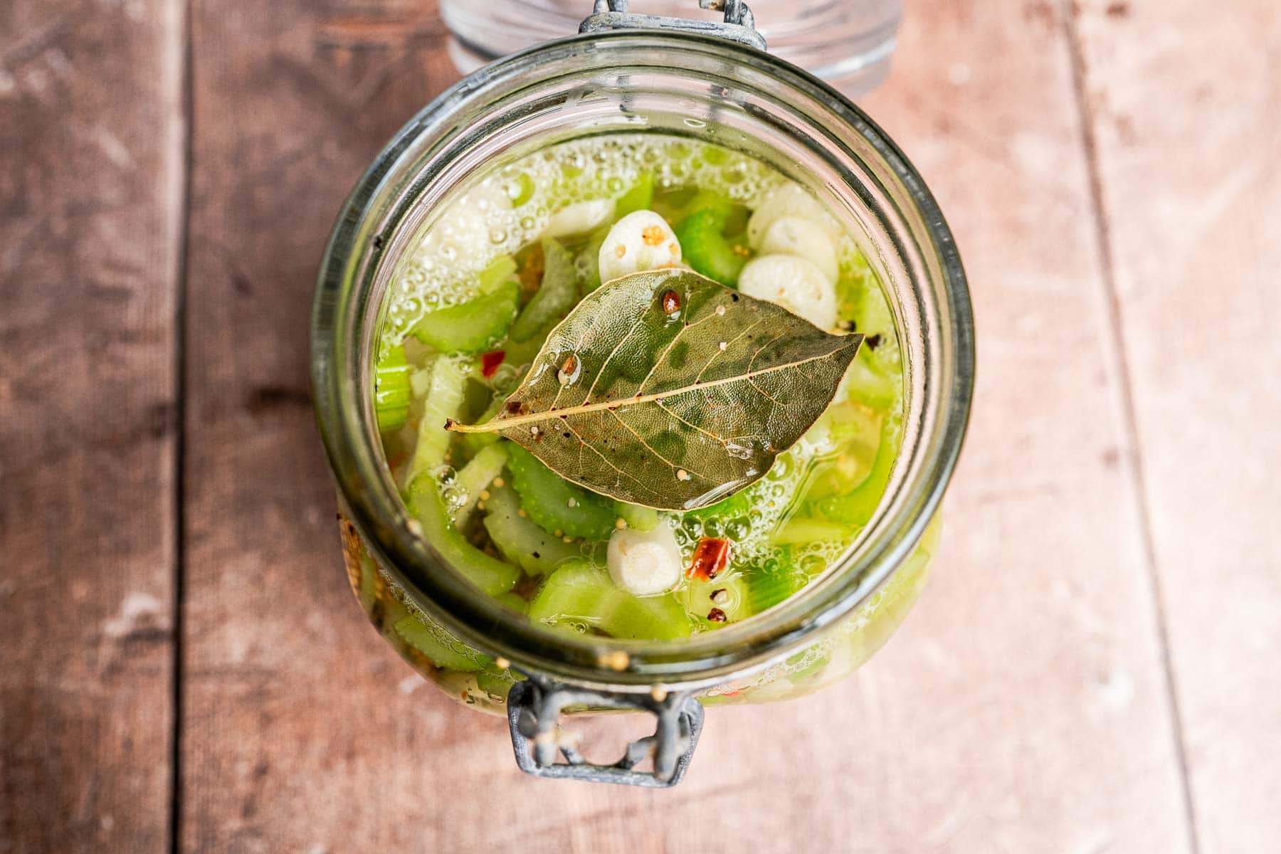 Open glass jar filled with pickled celery, sliced green vegetables, garlic cloves, and a bay leaf, sitting on a wooden surface.