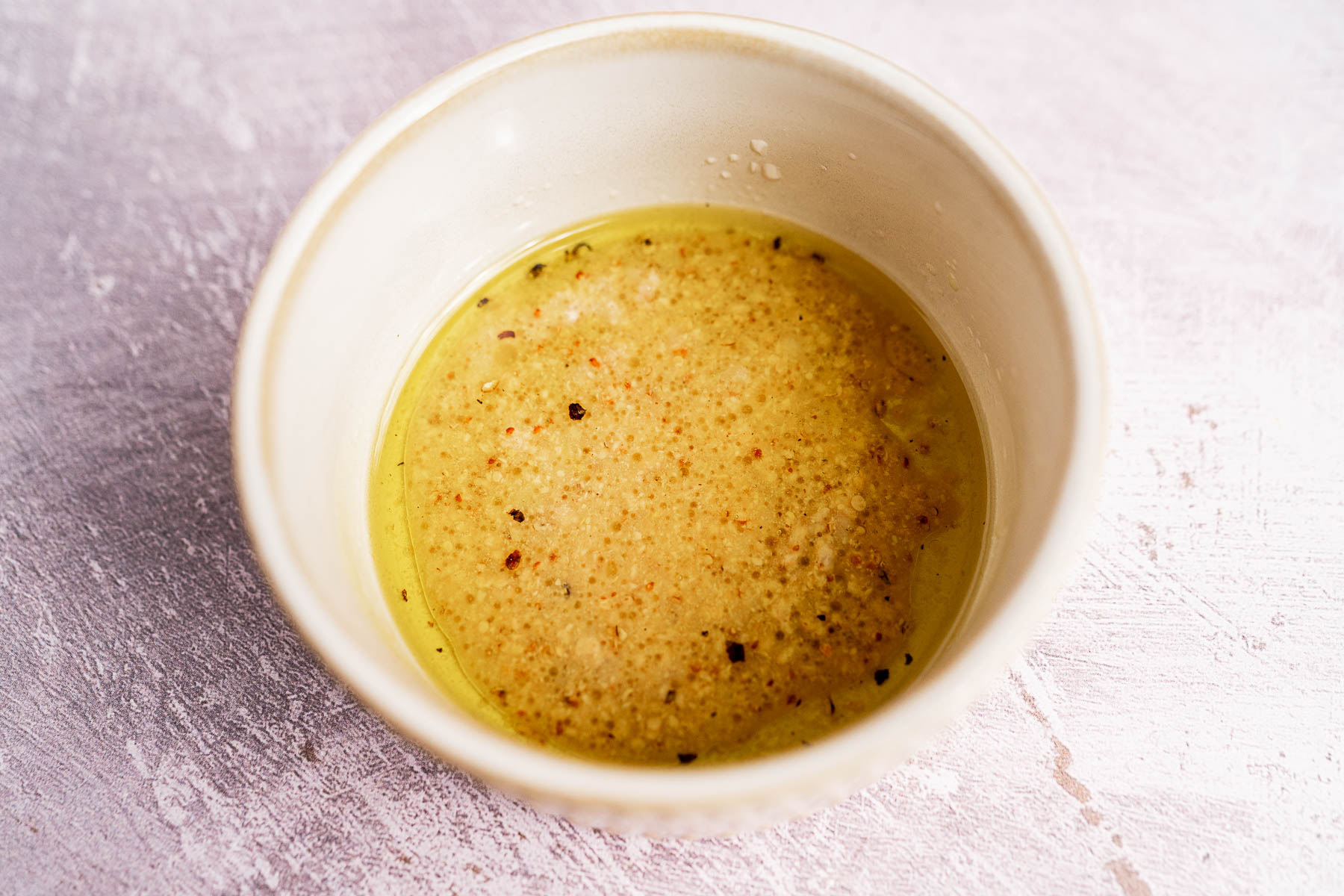 A white bowl containing a mixture of oil, vinegar, and visible seasonings sits on a light, textured surface—perfect for drizzling over a fresh spinach and arugula salad.