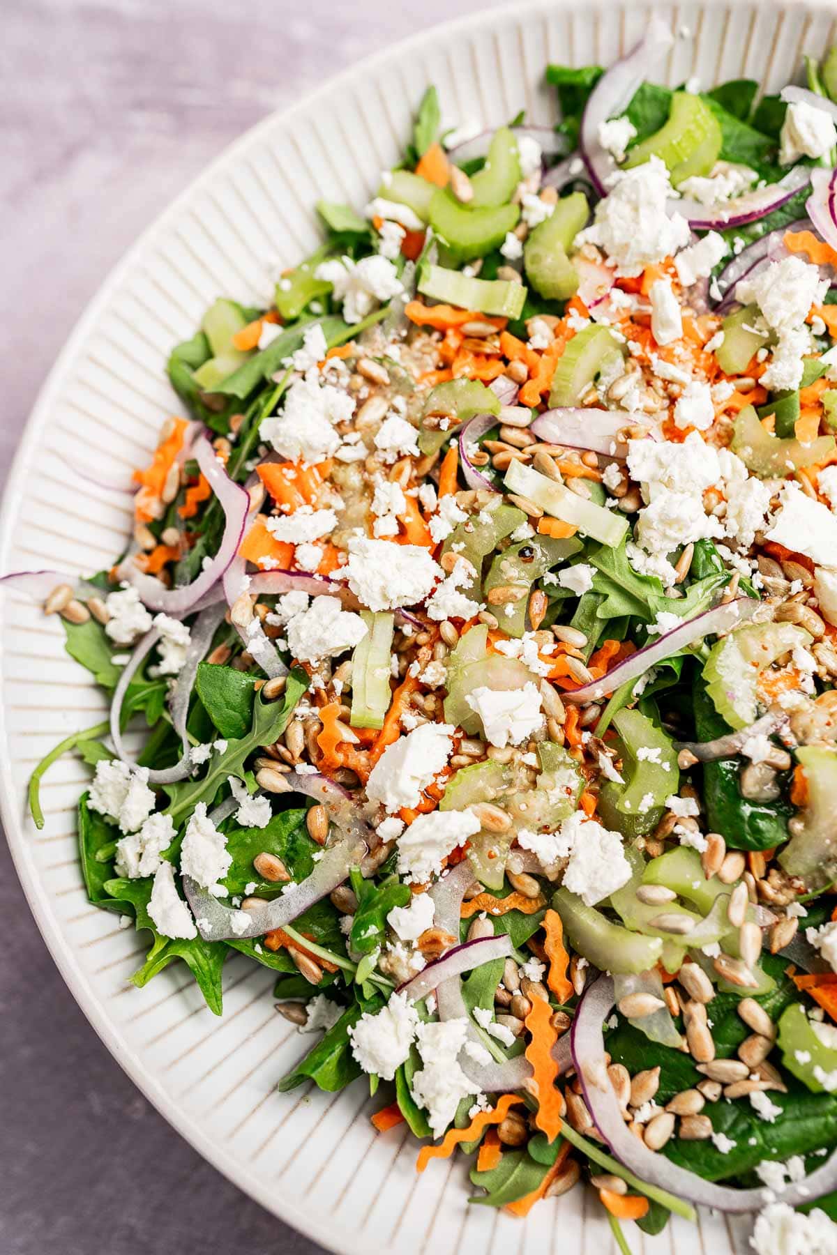 A close-up of a spinach and arugula salad with shredded carrots, sliced cucumber, red onion, feta cheese, sunflower seeds, and dressing in a white bowl.