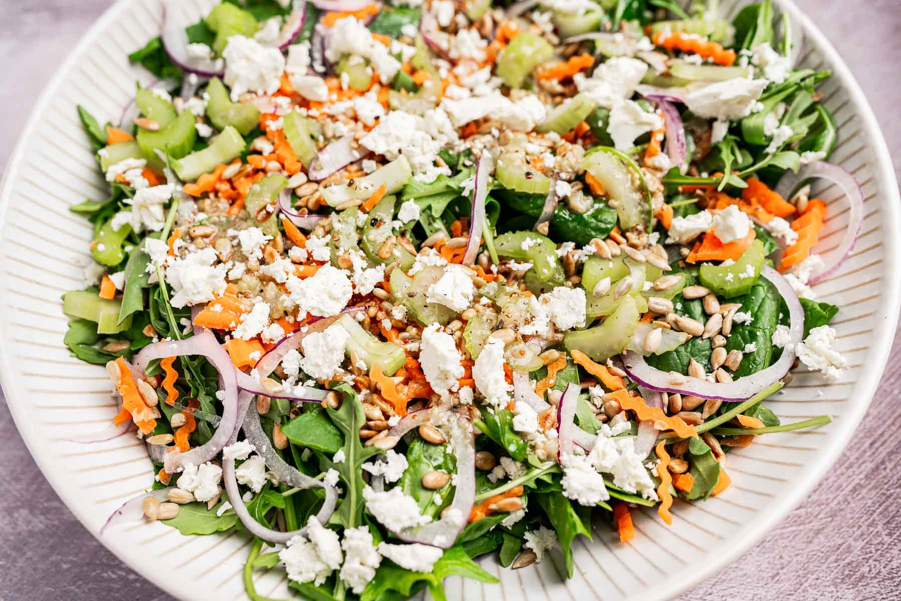 A large plate of mixed spinach and arugula salad topped with crumbled feta cheese, sunflower seeds, sliced red onions, grated carrots, and sliced celery.