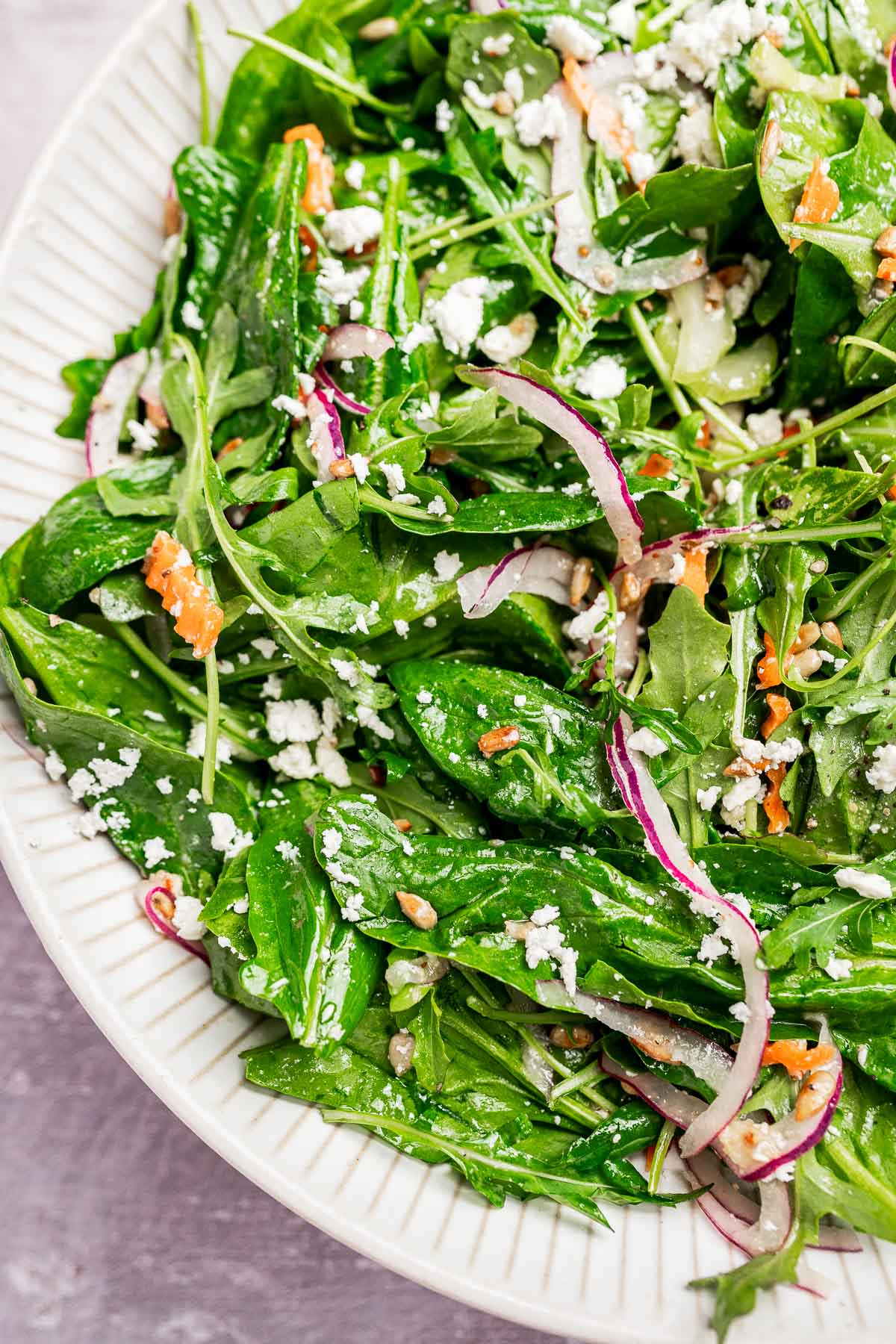 A close-up of a fresh spinach and arugula salad with red onion, shredded carrot, sunflower seeds, and crumbled feta cheese in a white bowl.