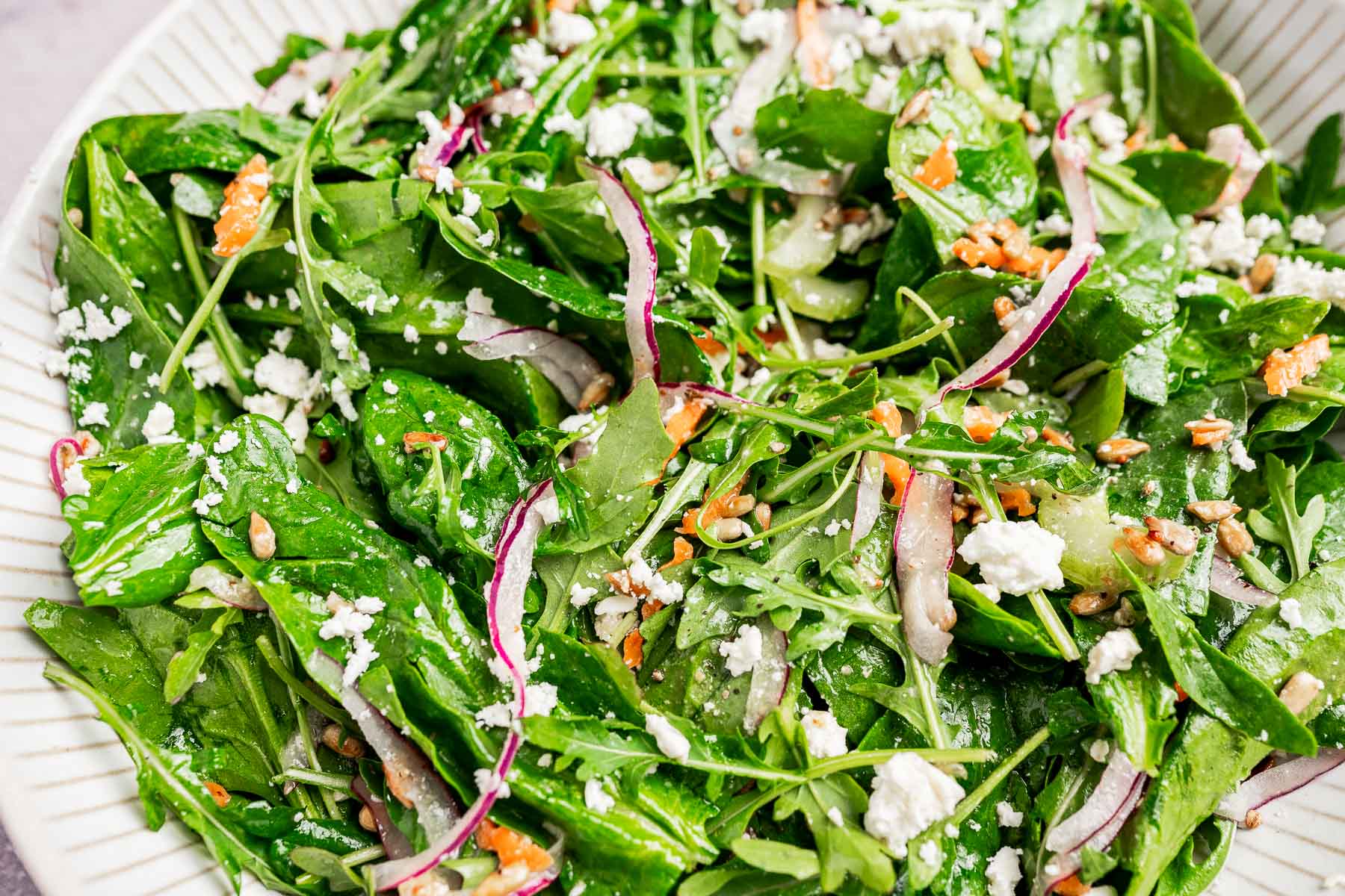 A close-up of a vibrant spinach and arugula salad, topped with red onion slices, crumbled feta cheese, and sunflower seeds on a white plate.