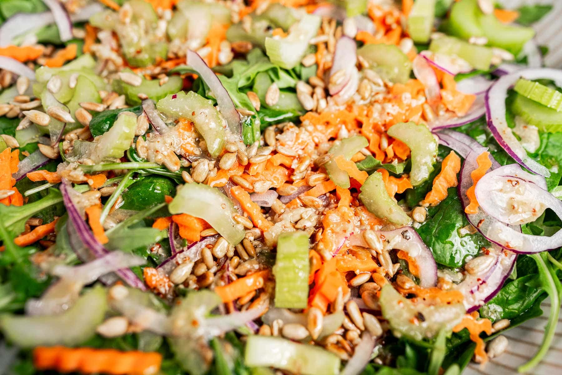 A close-up of a fresh spinach and arugula salad with shredded carrots, sliced celery, red onion, and sunflower seeds.