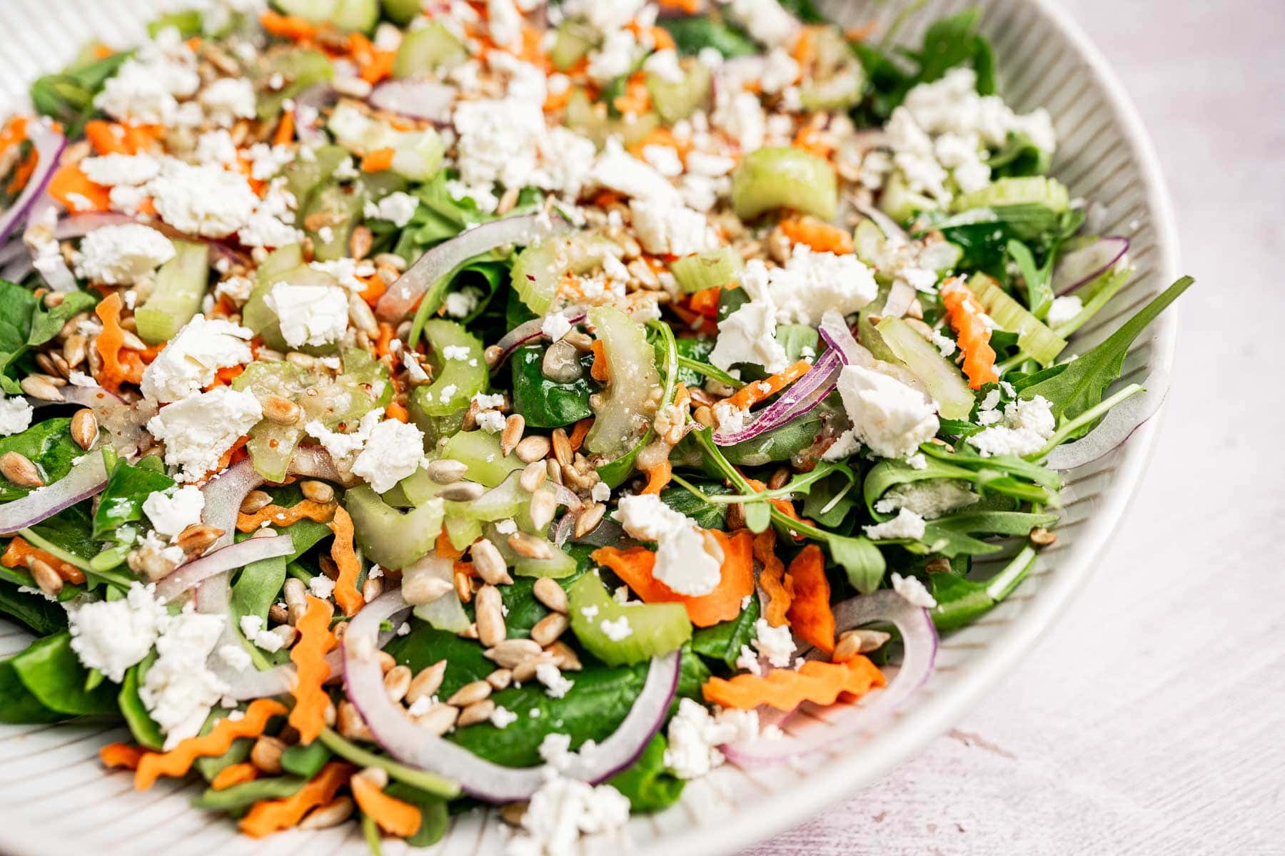 A close-up of a fresh spinach and arugula salad with mixed greens, sliced red onions, shredded carrots, celery, sunflower seeds, and crumbled feta cheese in a white bowl.