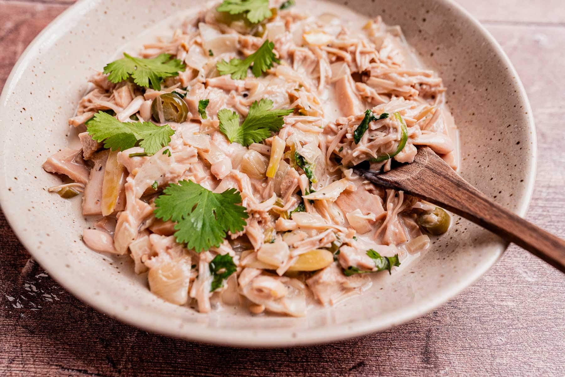 A ceramic bowl filled with ginataang langka—shredded jackfruit in a creamy sauce—garnished with fresh cilantro leaves, with a wooden spoon resting in the mixture.
