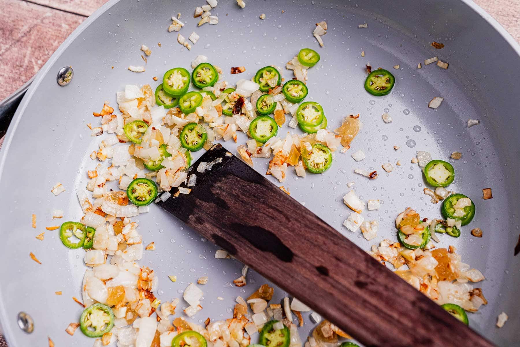 Chopped onions and sliced green chilies being sautéed in a frying pan with a wooden spatula, forming the flavorful base for ginataang langka.