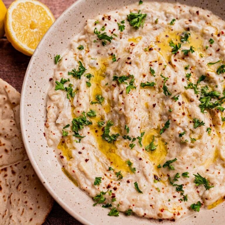 A bowl of baba ganoush, similar to mutabal, topped with chopped parsley, olive oil, and red pepper flakes, served with pita bread and a halved lemon on the side.