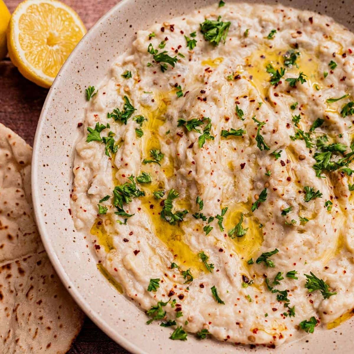 A bowl of baba ganoush, similar to mutabal, topped with chopped parsley, olive oil, and red pepper flakes, served with pita bread and a halved lemon on the side.