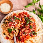 A plate of spaghetti topped with chunky vegetarian bolognese sauce, grated cheese, and fresh parsley, with a fork and a small bowl of grated cheese on the side.