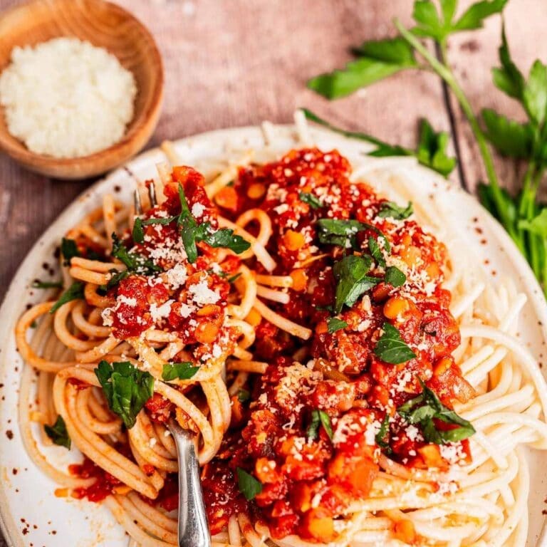 A plate of spaghetti topped with chunky vegetarian bolognese sauce, grated cheese, and fresh parsley, with a fork and a small bowl of grated cheese on the side.