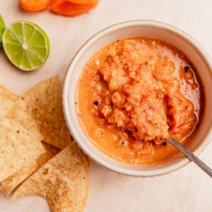A bowl of vibrant habanero salsa with a spoon, surrounded by tortilla chips, lime halves, and an orange pepper on a light surface.
