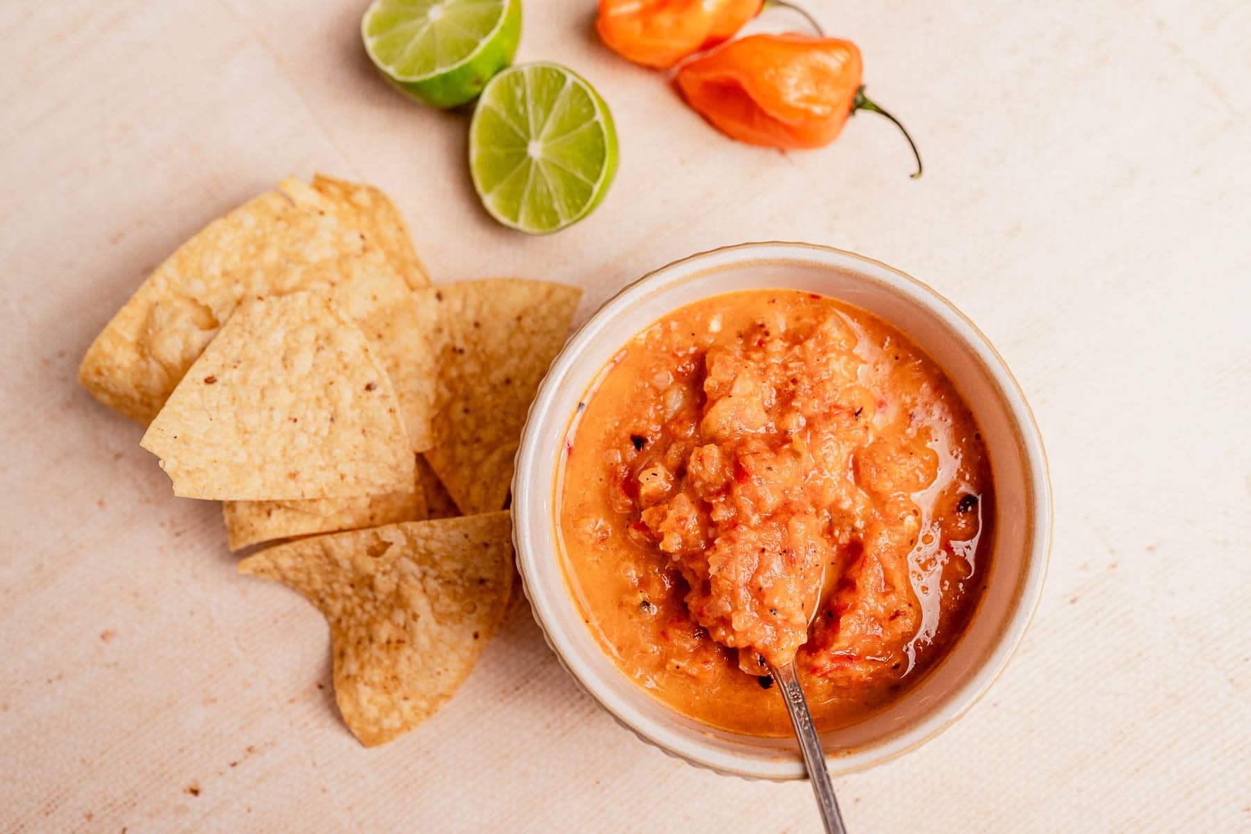 A bowl of vibrant habanero salsa with a spoon is surrounded by tortilla chips, halved limes, and whole habanero peppers on a light surface.