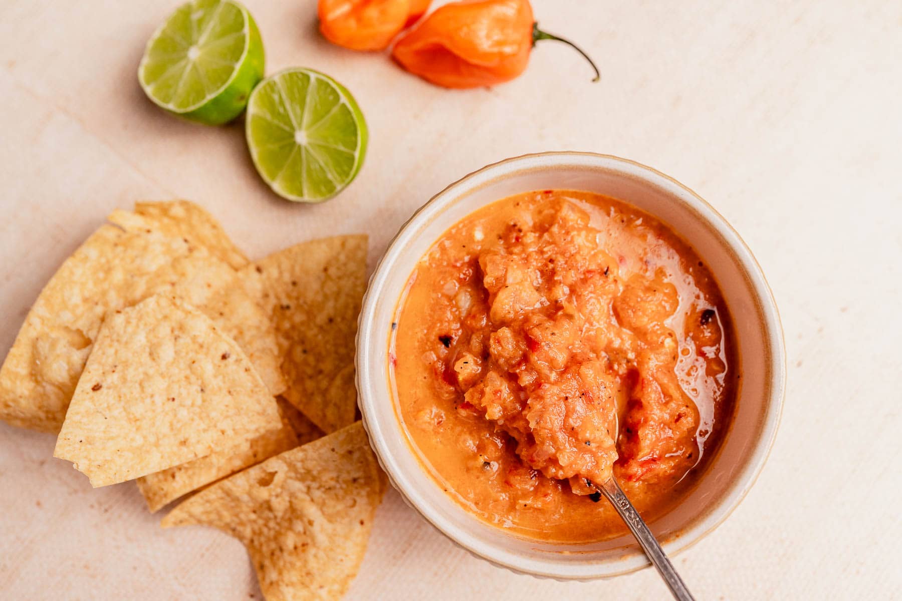 A bowl of vibrant habanero salsa with a spoon, surrounded by tortilla chips, halved limes, and a whole habanero pepper on a light surface.