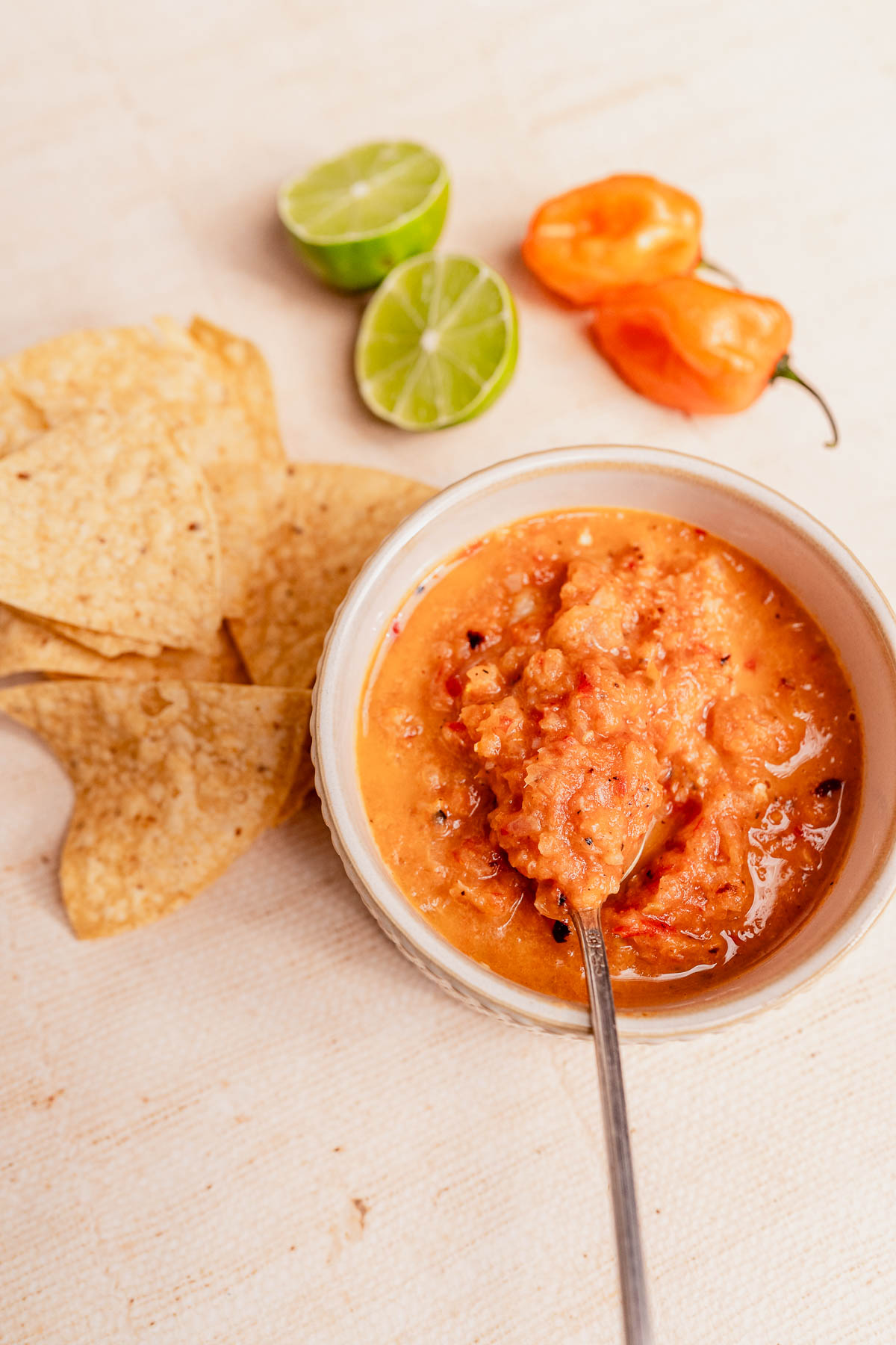 A bowl of chunky habanero salsa with a spoon, surrounded by tortilla chips, lime halves, and a habanero pepper on a light surface.