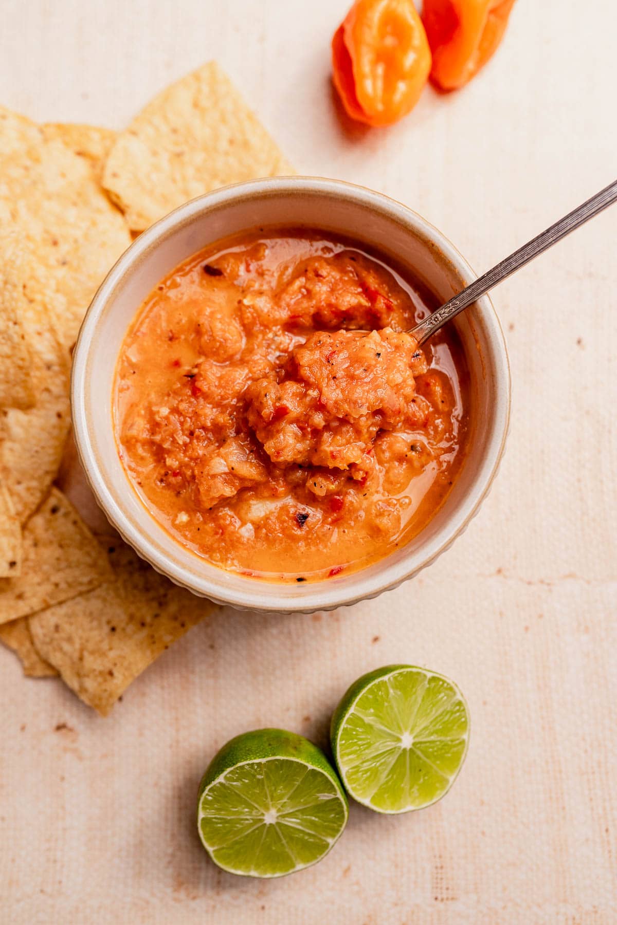 A bowl of vibrant habanero salsa with a spoon, surrounded by tortilla chips, two lime halves, and an orange pepper on a light surface.