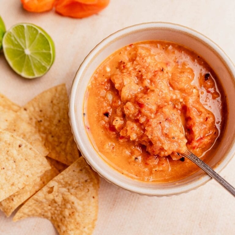 A bowl of vibrant habanero salsa with a spoon, surrounded by tortilla chips, lime halves, and an orange pepper on a light surface.