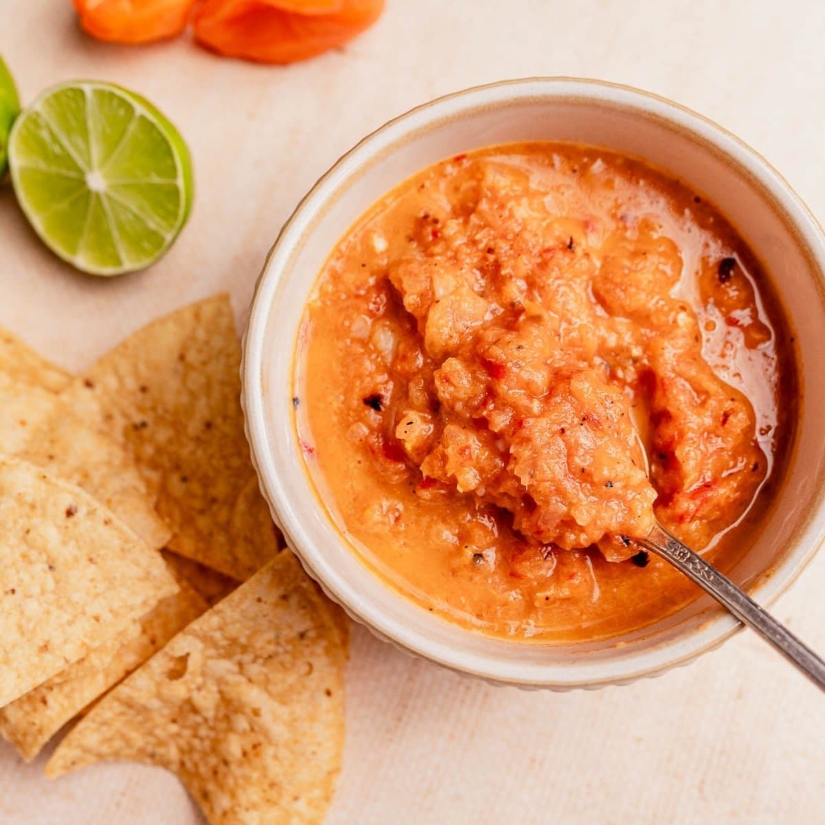 A bowl of vibrant habanero salsa with a spoon, surrounded by tortilla chips, lime halves, and an orange pepper on a light surface.