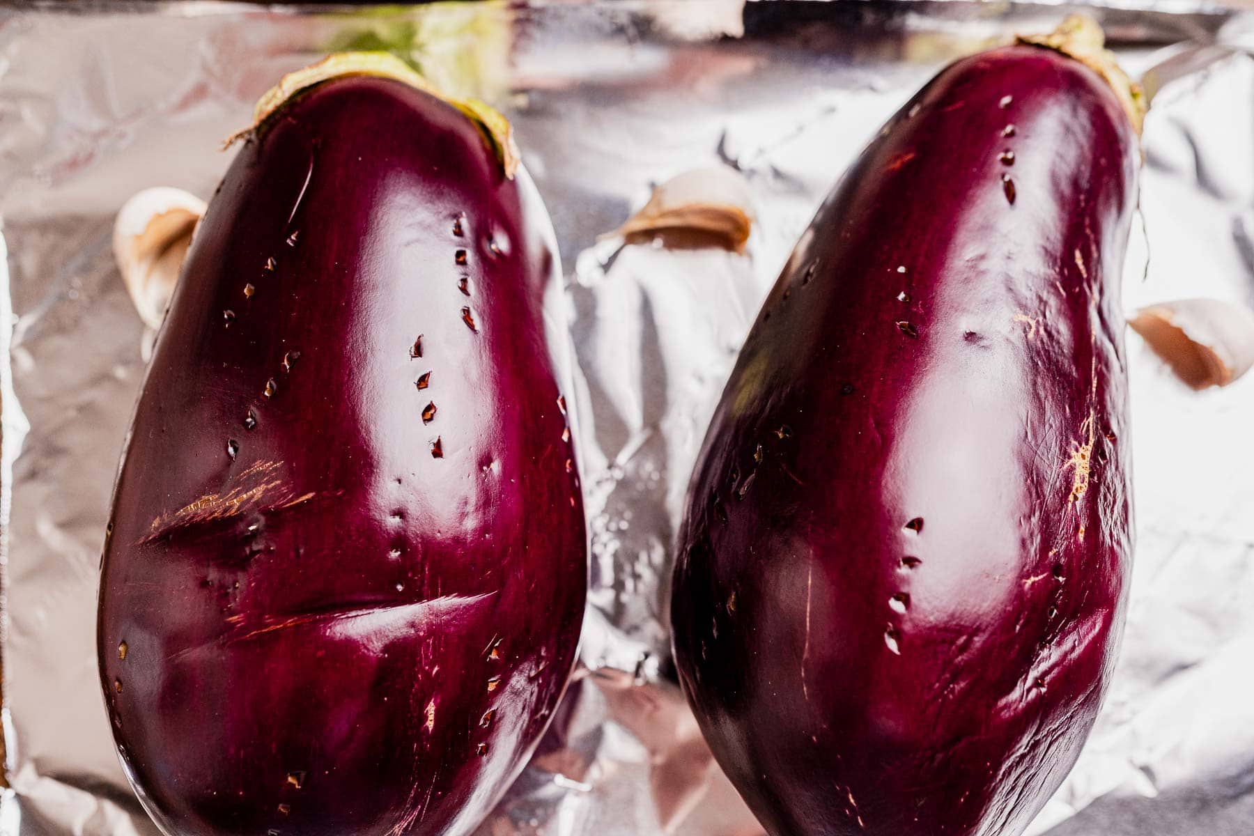 Two whole eggplants with pricked skin rest on a sheet of aluminum foil, accompanied by a few garlic cloves—perfect for roasting and making smoky mutabal.