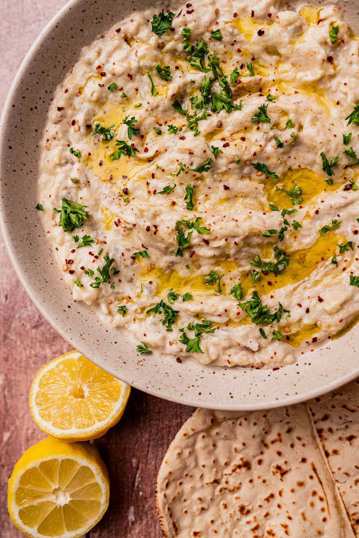 A bowl of baba ganoush, similar to mutabal, garnished with chopped parsley and olive oil, with lemon halves and flatbread on the side.