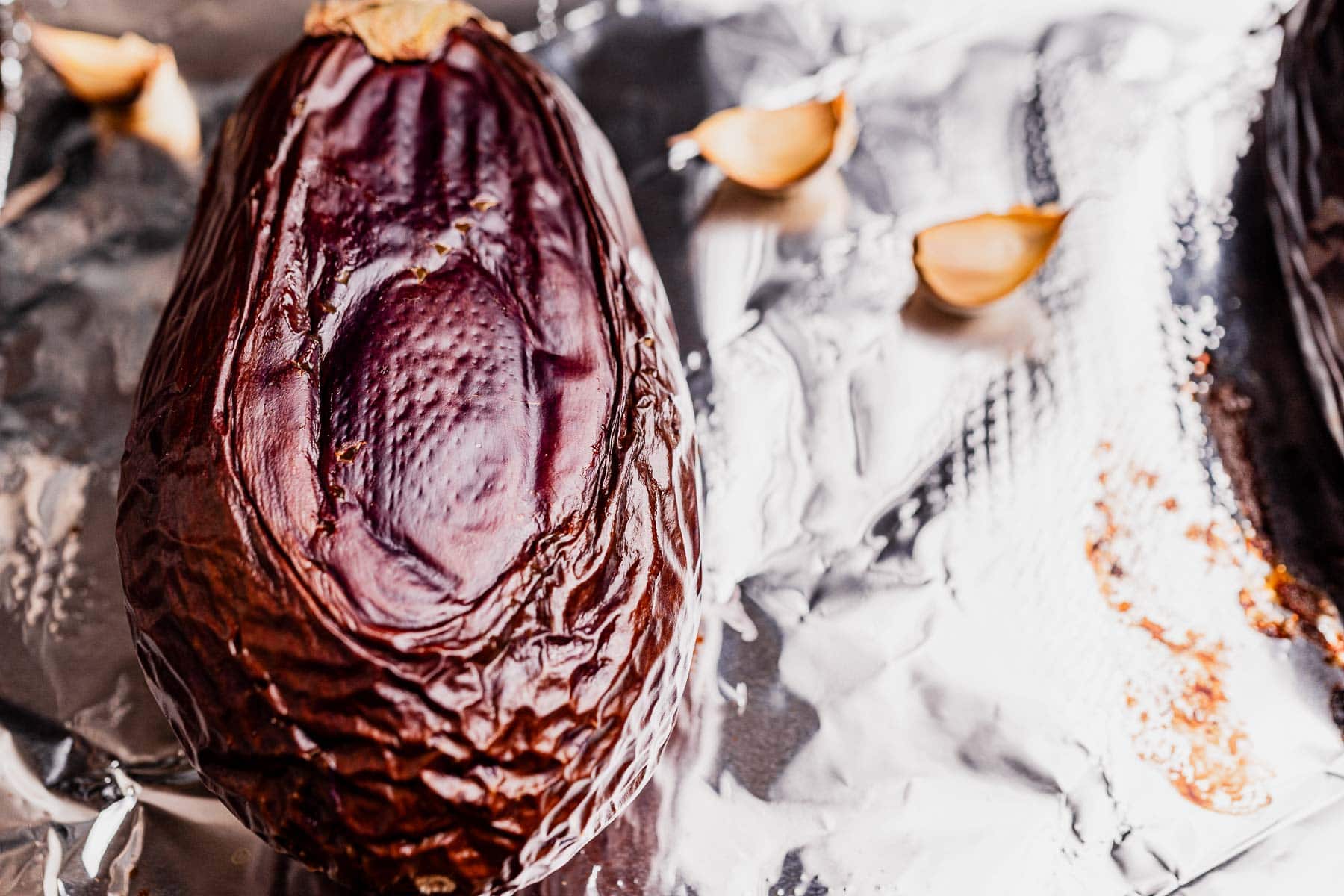 A roasted eggplant half with wrinkled skin, perfect for making mutabal, rests on aluminum foil next to cloves of garlic.