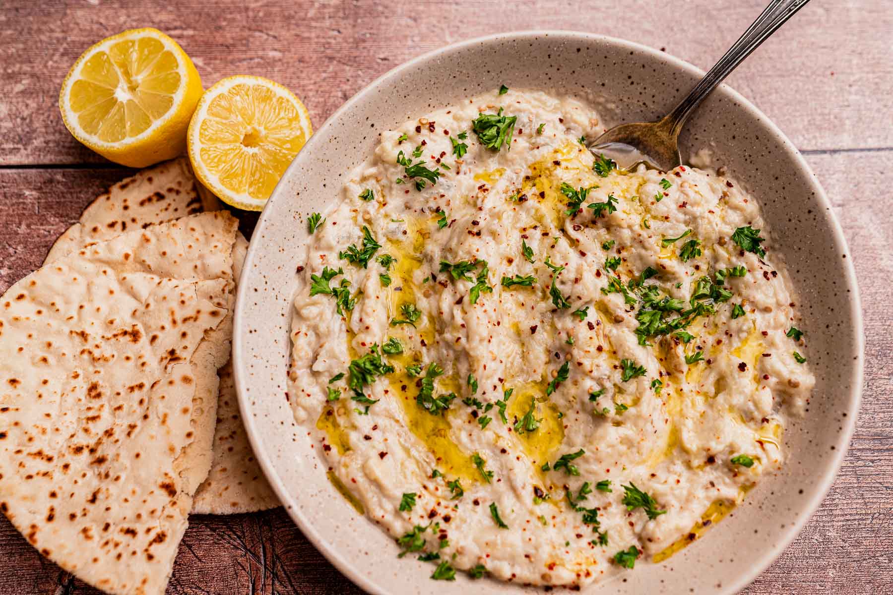 A bowl of baba ganoush, similar to mutabal, garnished with parsley and olive oil, served with flatbread and lemon halves on a wooden surface.