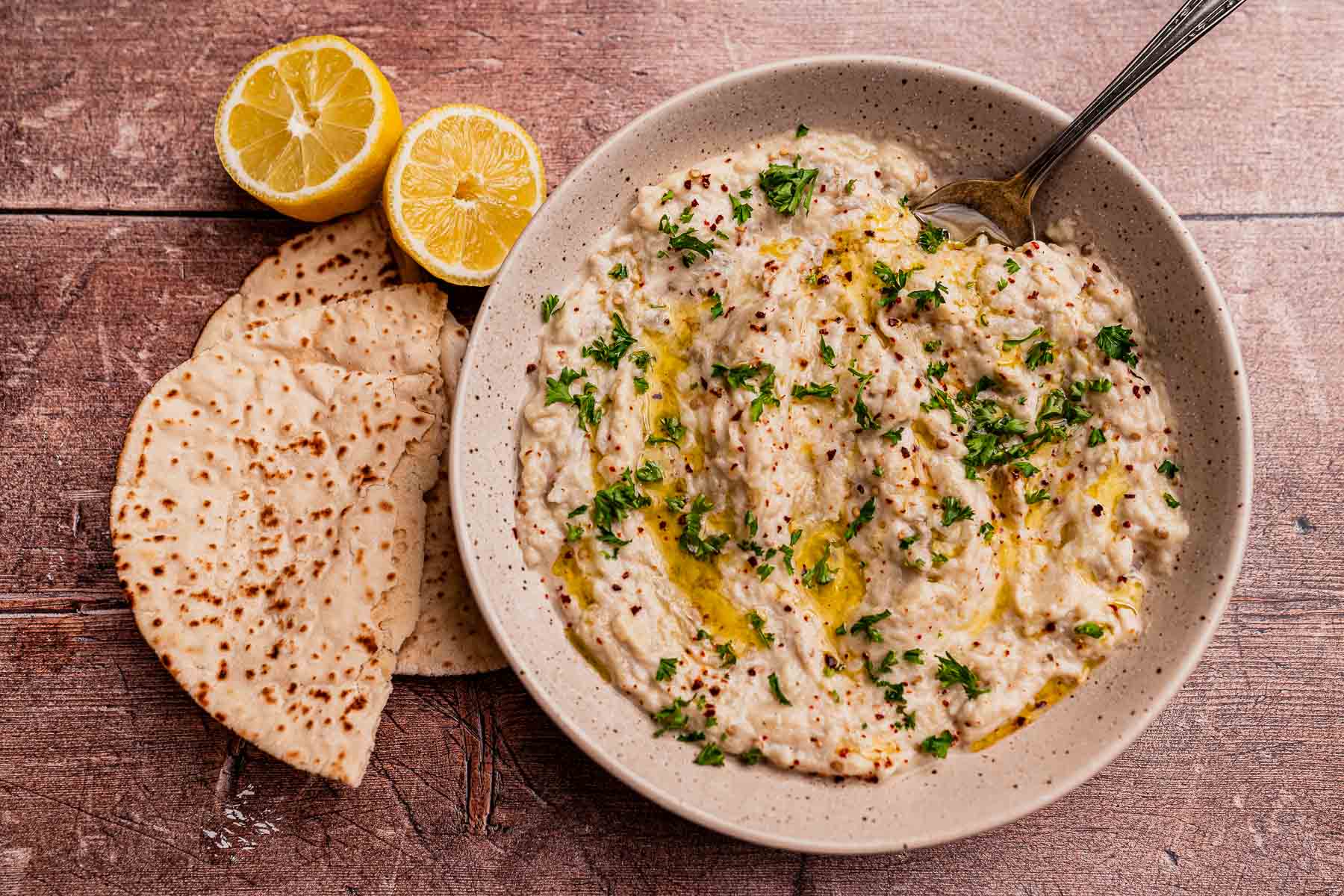 A bowl of baba ganoush, similar to mutabal, garnished with parsley and olive oil, served with lemon halves and pita bread on a wooden surface.