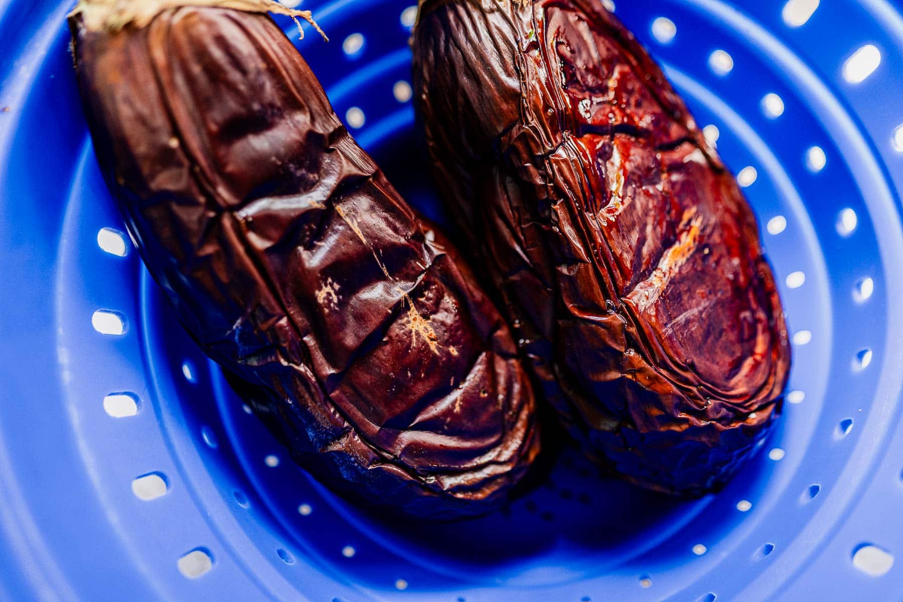 Two roasted eggplants with wrinkled skins, perfect for making mutabal, are placed in a blue plastic colander with holes.
