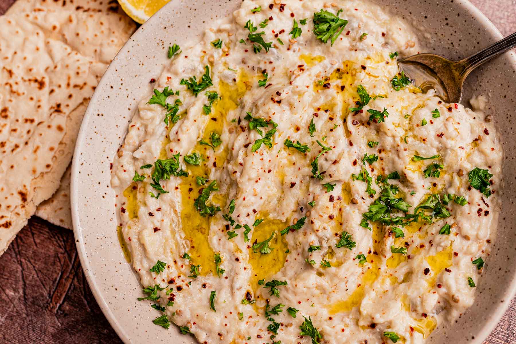 Bowl of creamy baba ganoush, similar to mutabal, topped with chopped parsley and olive oil, served with a spoon and slices of flatbread on the side.