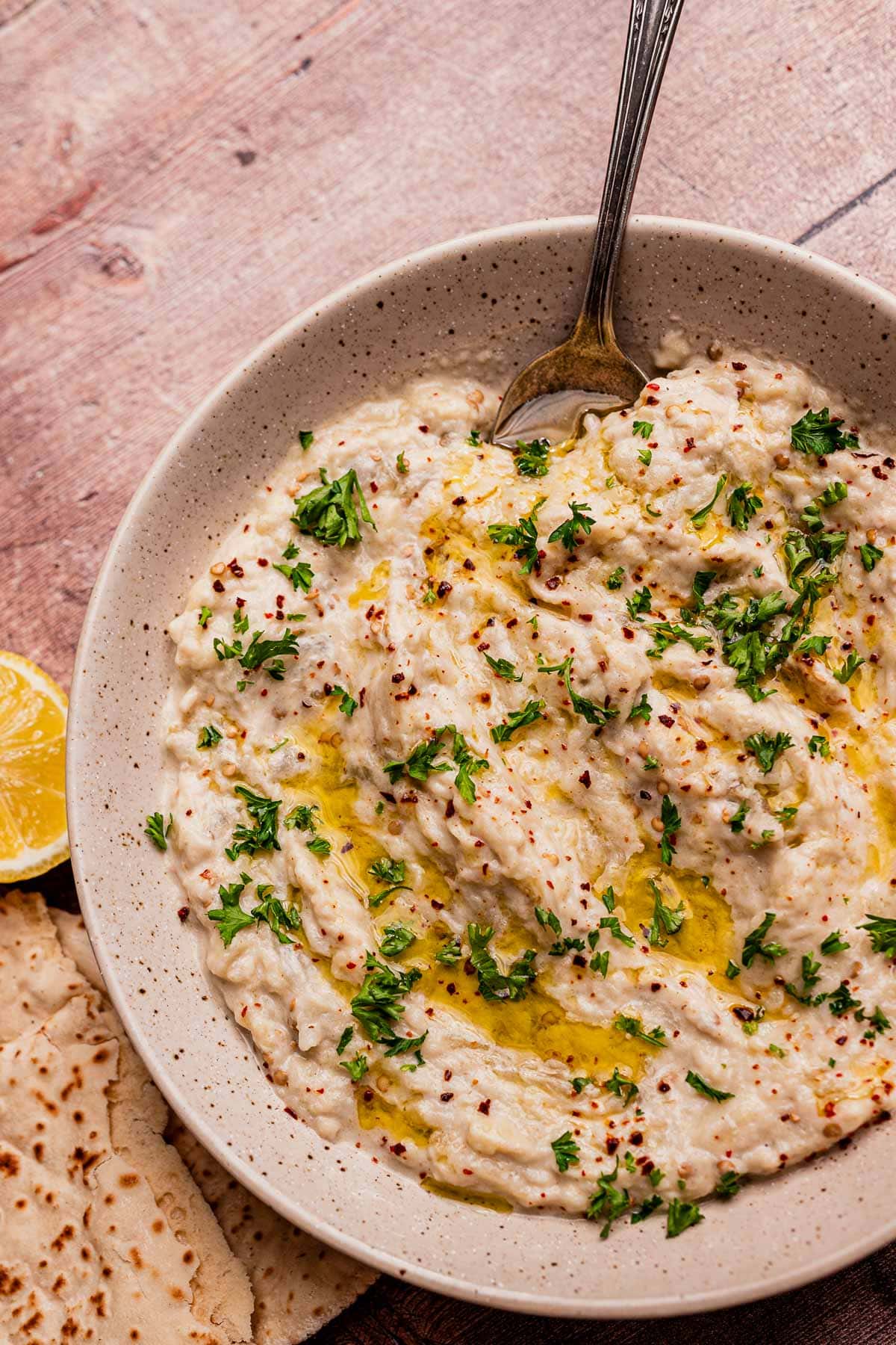 A bowl of creamy baba ganoush, similar to mutabal, topped with olive oil, chopped parsley, and spices, with a spoon in the bowl and flatbread and a lemon wedge on the side.