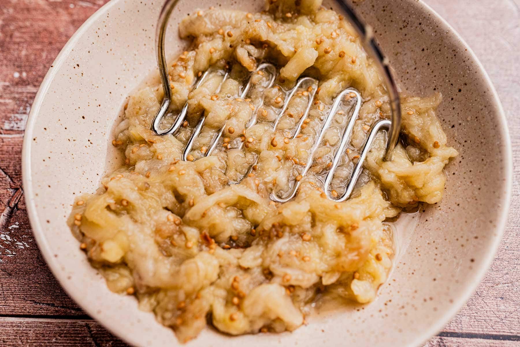 A bowl of mashed roasted eggplant, perfect for mutabal, being pressed with a metal potato masher on a wooden surface.