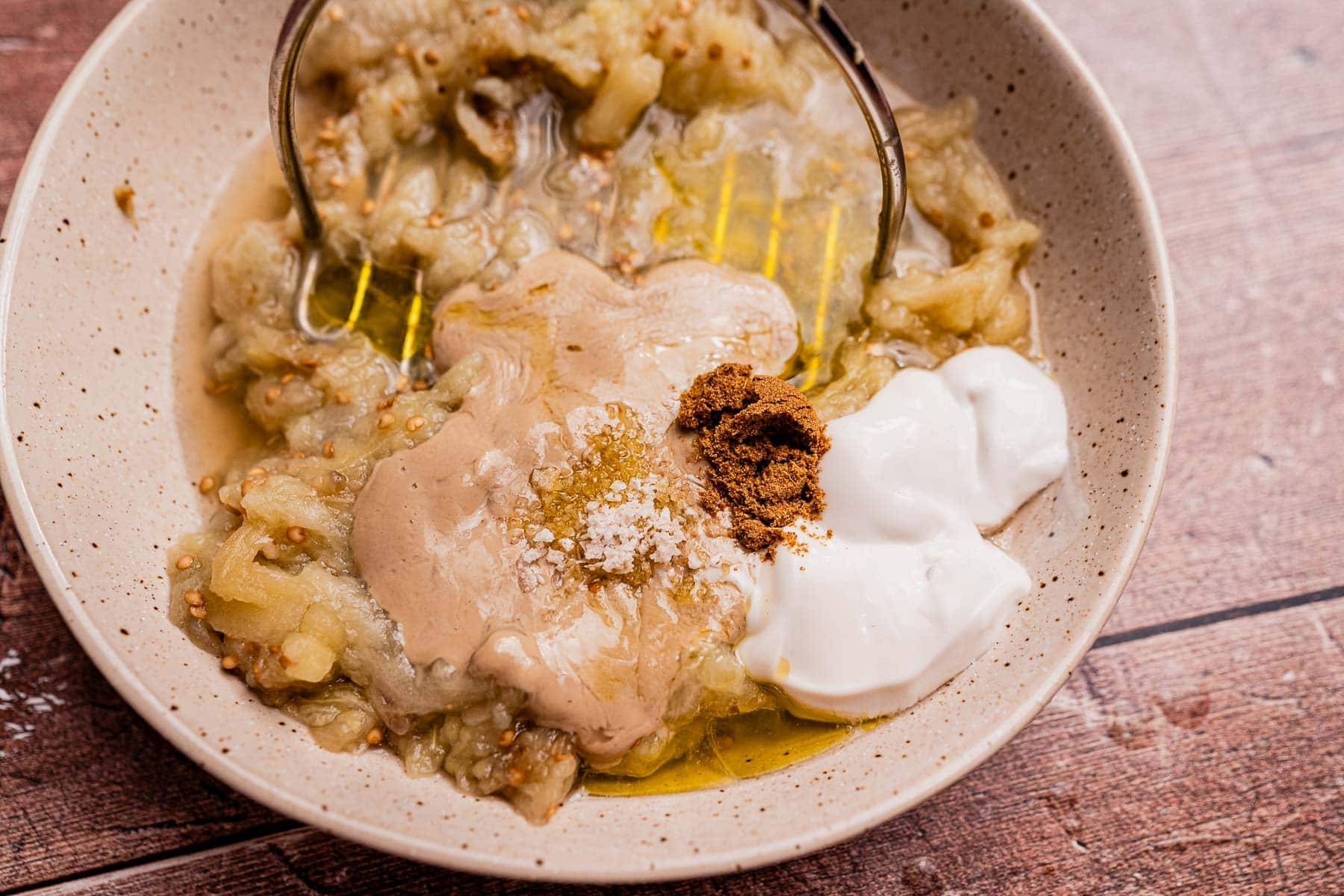 A bowl of mutabal with mashed eggplant, tahini, yogurt, ground spices, olive oil, and salt sits ready to be mixed on a wooden surface.