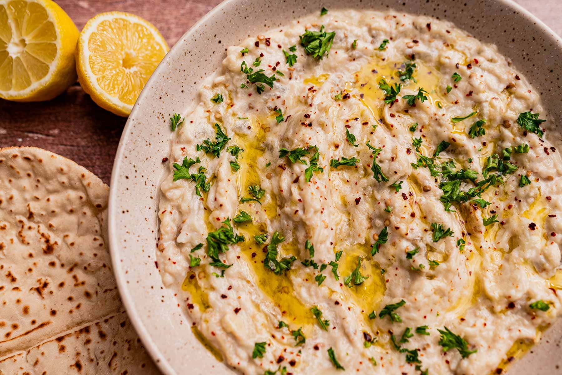 A bowl of baba ganoush, similar to mutabal, topped with chopped parsley, olive oil, and spices sits next to flatbread and halved lemons on a wooden surface.