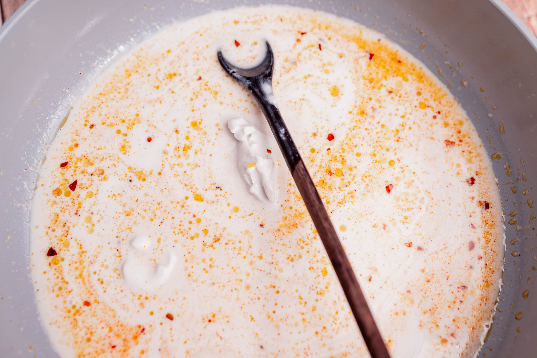 A wooden spoon rests in a pan of creamy coconut milk pasta sauce with orange oil and red pepper flakes swirling on the surface.