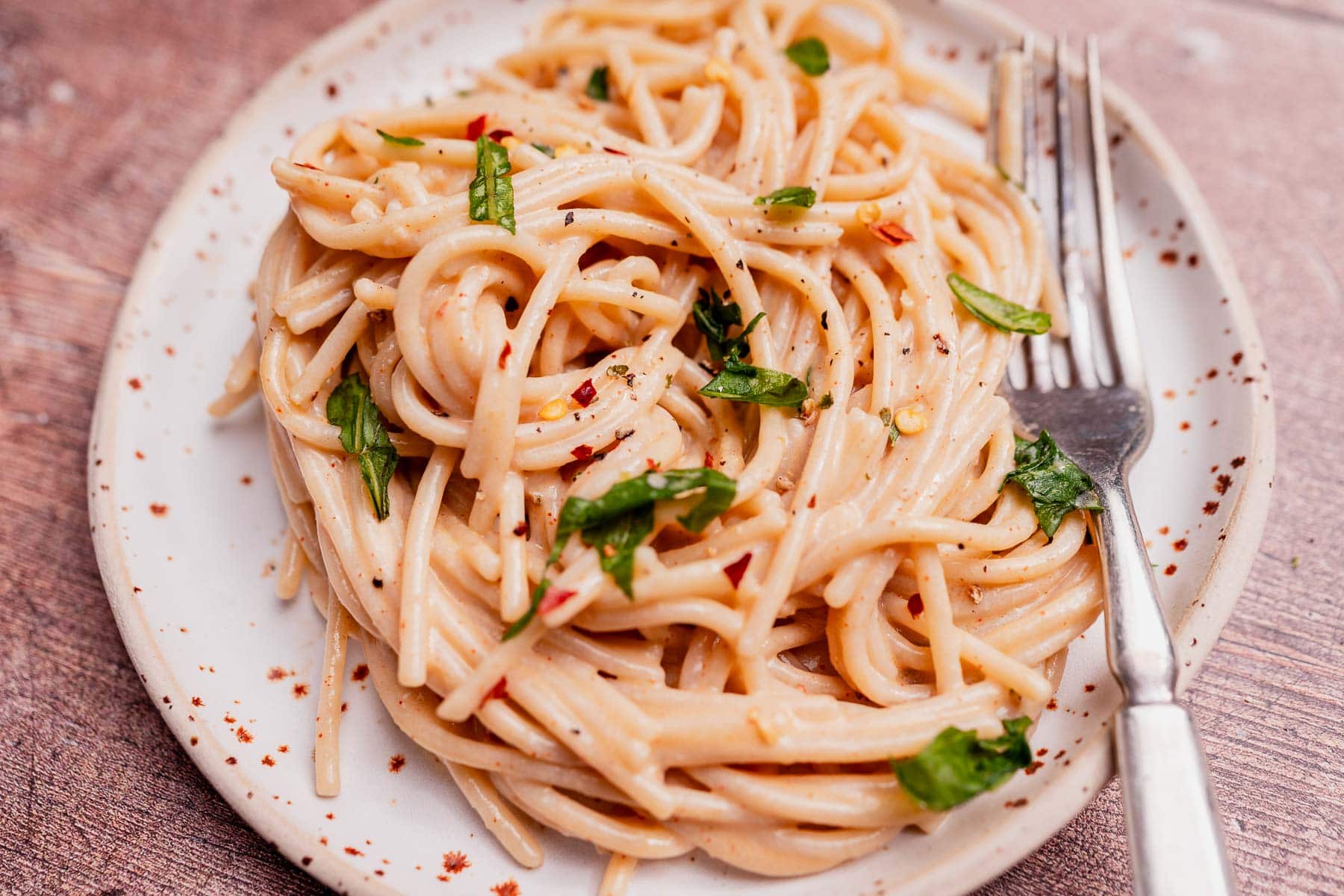 A plate of cooked spaghetti topped with creamy coconut milk pasta sauce, chopped herbs, and red pepper flakes, with a fork resting on the side.