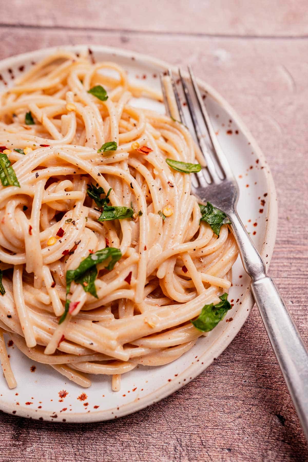 A plate of cooked spaghetti with creamy coconut milk pasta sauce, garnished with fresh herbs and red pepper flakes, sits next to a fork on a rustic wooden surface.