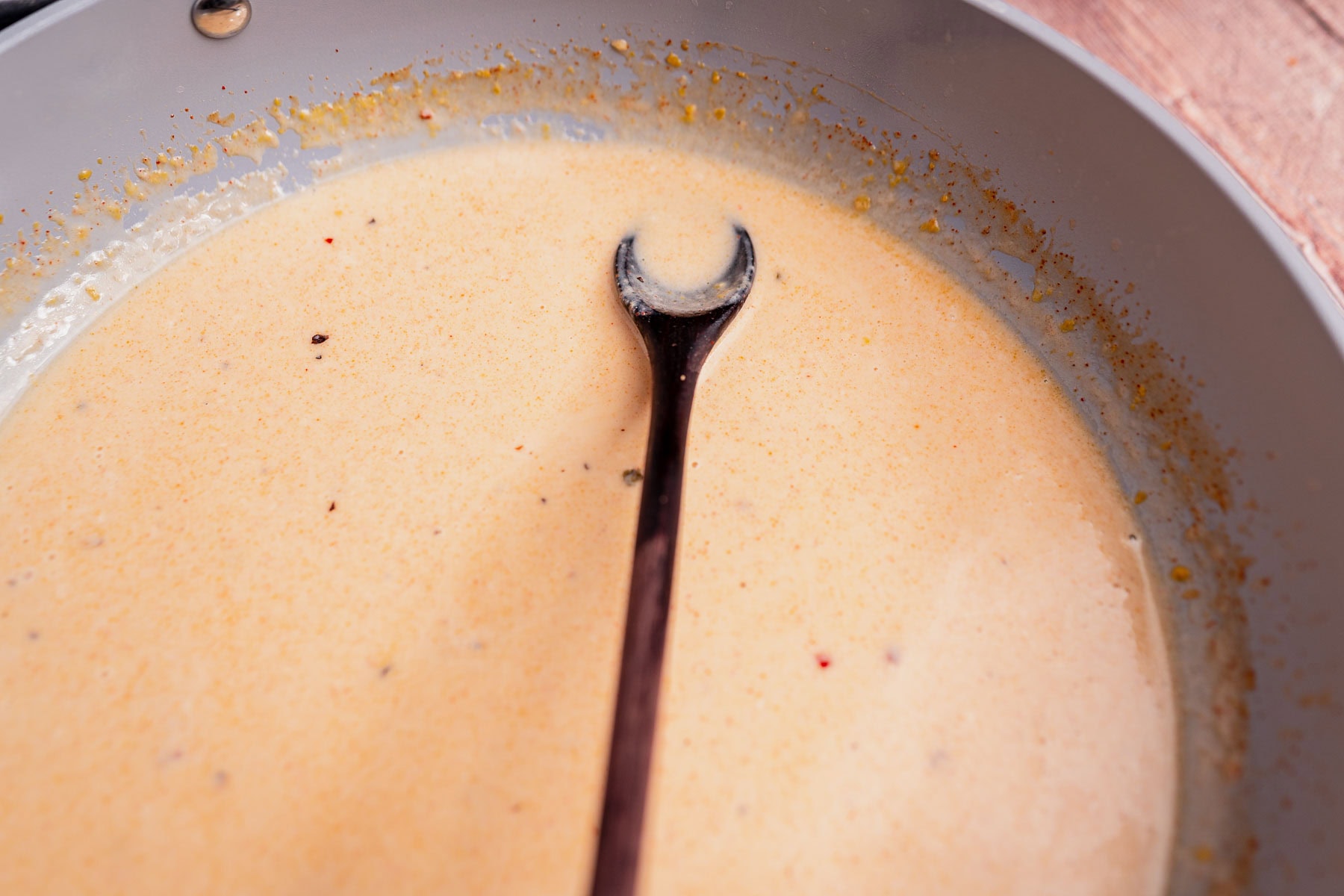 A wooden spoon rests in a pan filled with light brown coconut milk pasta sauce, with seasoning visible around the edges of the pan.
