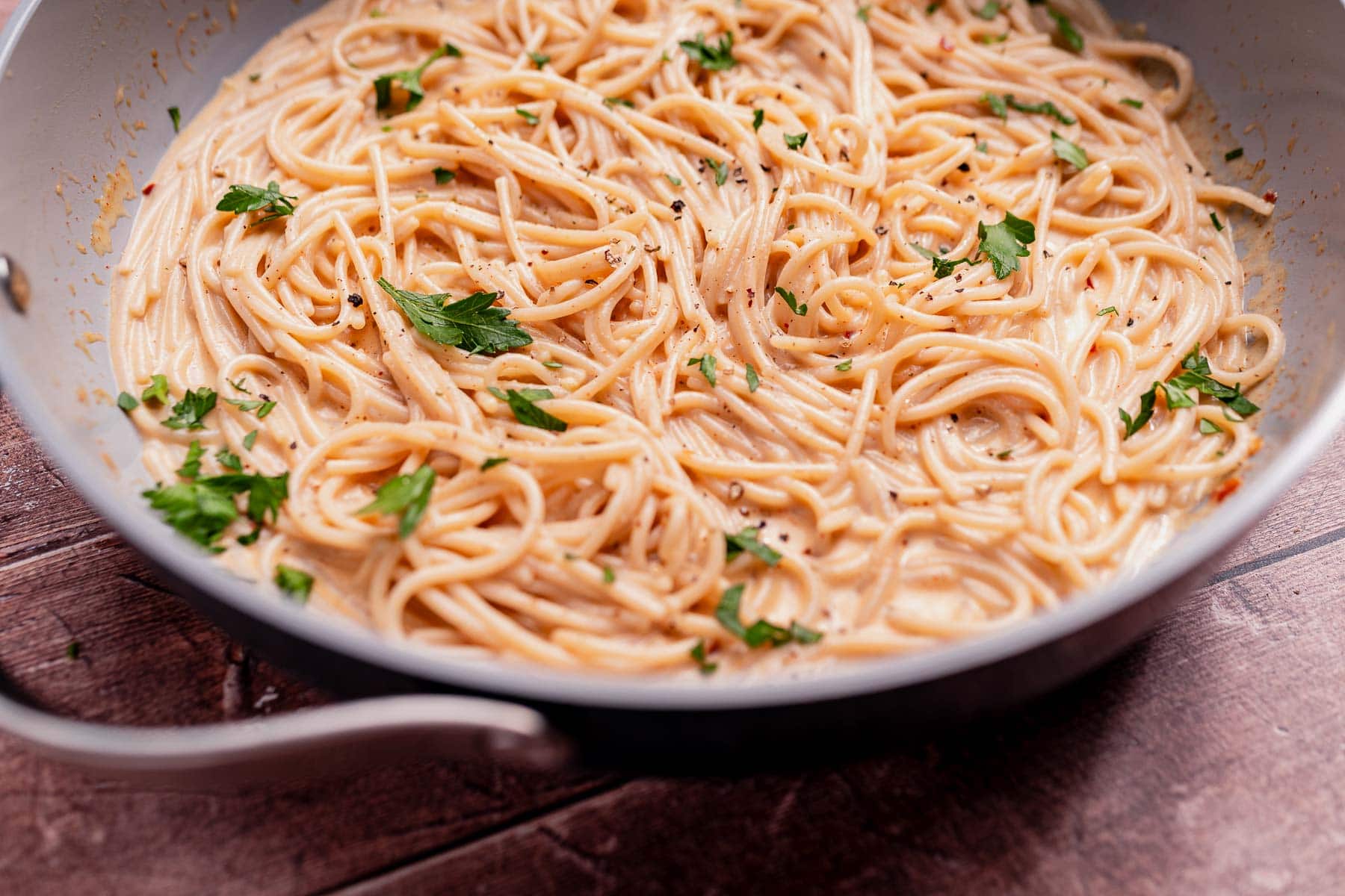 A pan filled with cooked spaghetti in a creamy coconut milk pasta sauce, garnished with chopped parsley and cracked black pepper, sits on a wooden surface.