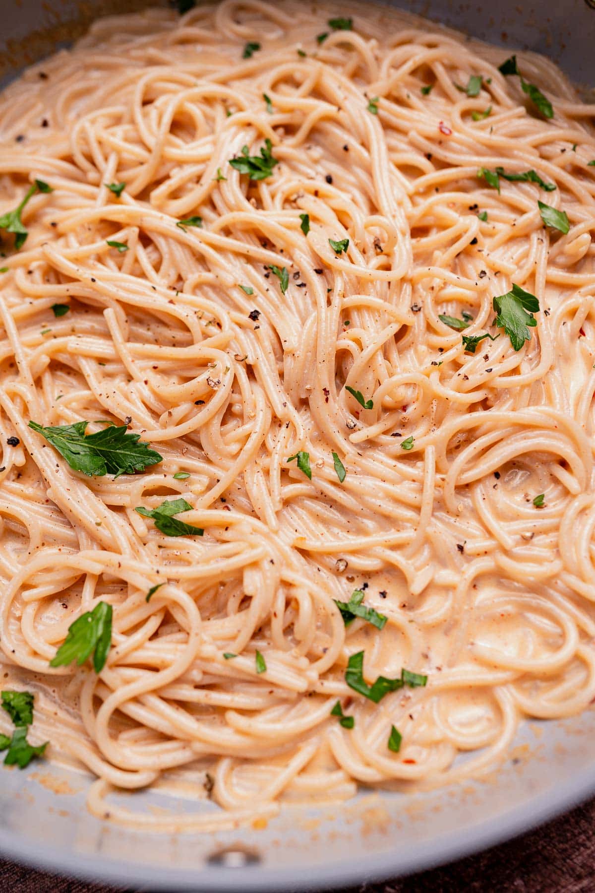 Cooked spaghetti in a creamy coconut milk pasta sauce, garnished with chopped parsley and black pepper, served in a shallow pan.