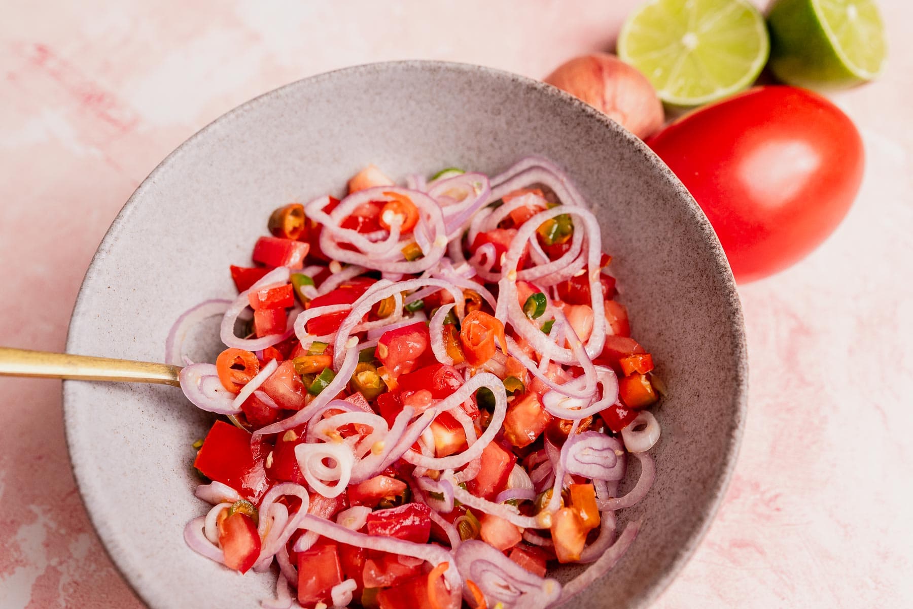 A bowl of sambal dabu dabu made with chopped tomatoes, sliced onions, and green chilies sits with a spoon on a light pink surface, beside a tomato, lime halves, and a shallot.