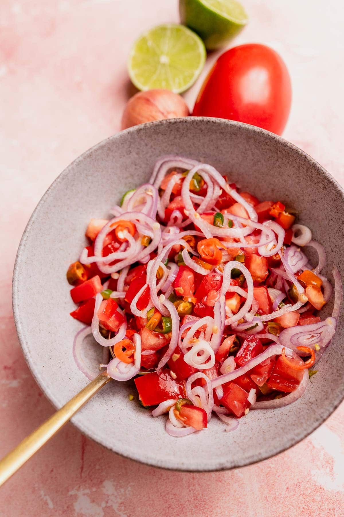 A gray bowl filled with sambal dabu dabu—a salad of chopped tomatoes, sliced red onions, and green chili, with a spoon. A halved lime, whole tomato, and shallot sit in the background.