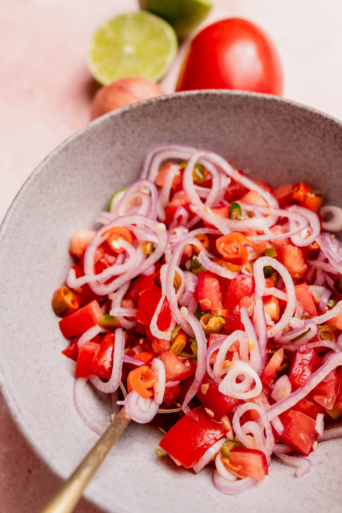 A bowl of salad inspired by sambal dabu dabu with sliced red onions, diced tomatoes, and green chilies, served with a spoon, and fresh lime and tomato in the background.