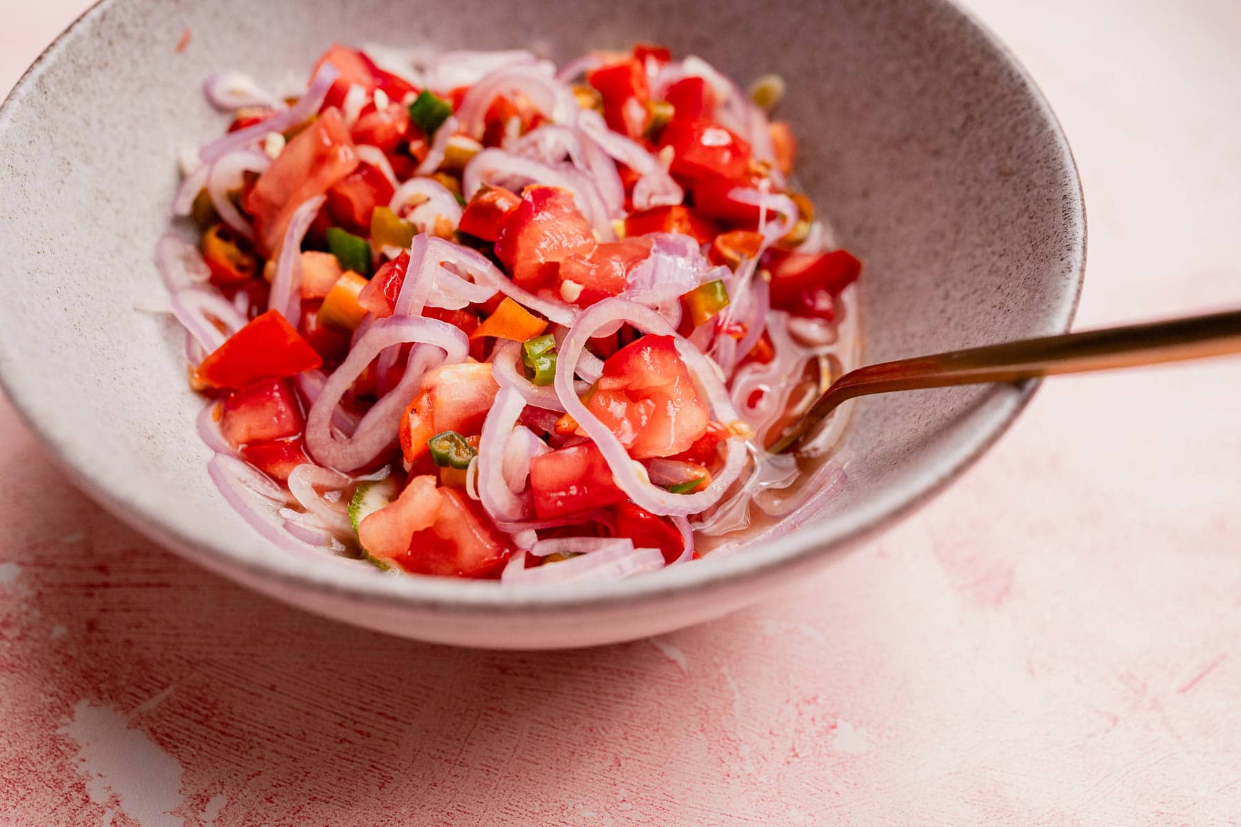A bowl filled with sambal dabu dabu salad, featuring sliced onions, diced tomatoes, green chilies, and other vegetables, with a fork resting in the bowl.