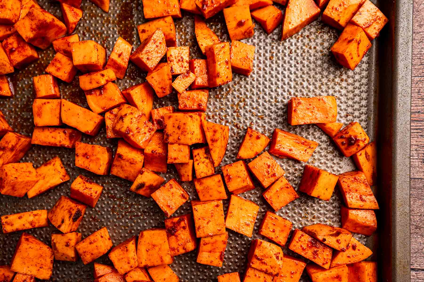Cubed sweet potatoes coated in seasoning are spread out on a textured metal baking sheet, ready for roasting into a flavorful sweet potato hash.