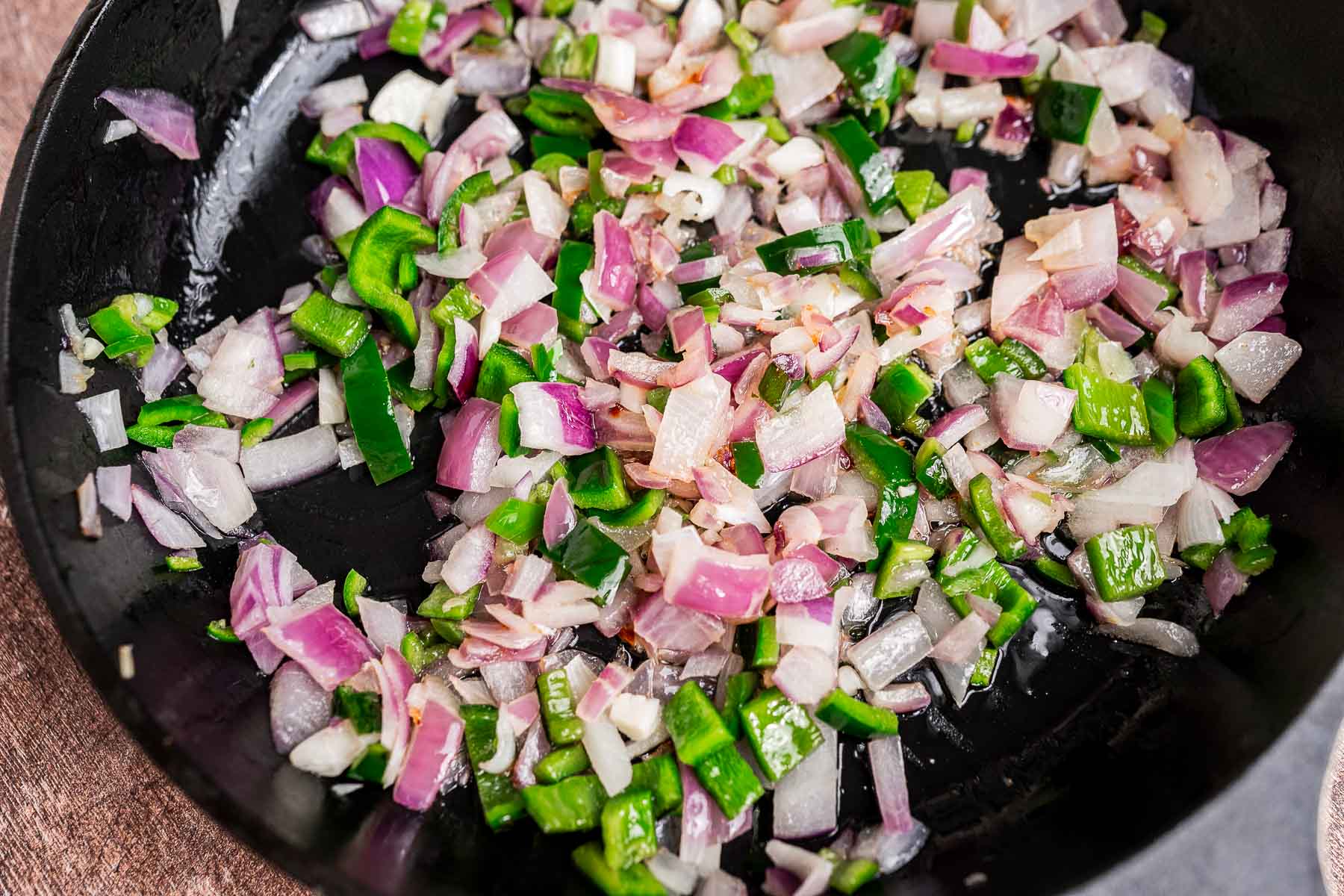 Chopped red onions and green bell peppers are being sautéed in a black skillet as the flavorful base for a sweet potato hash.