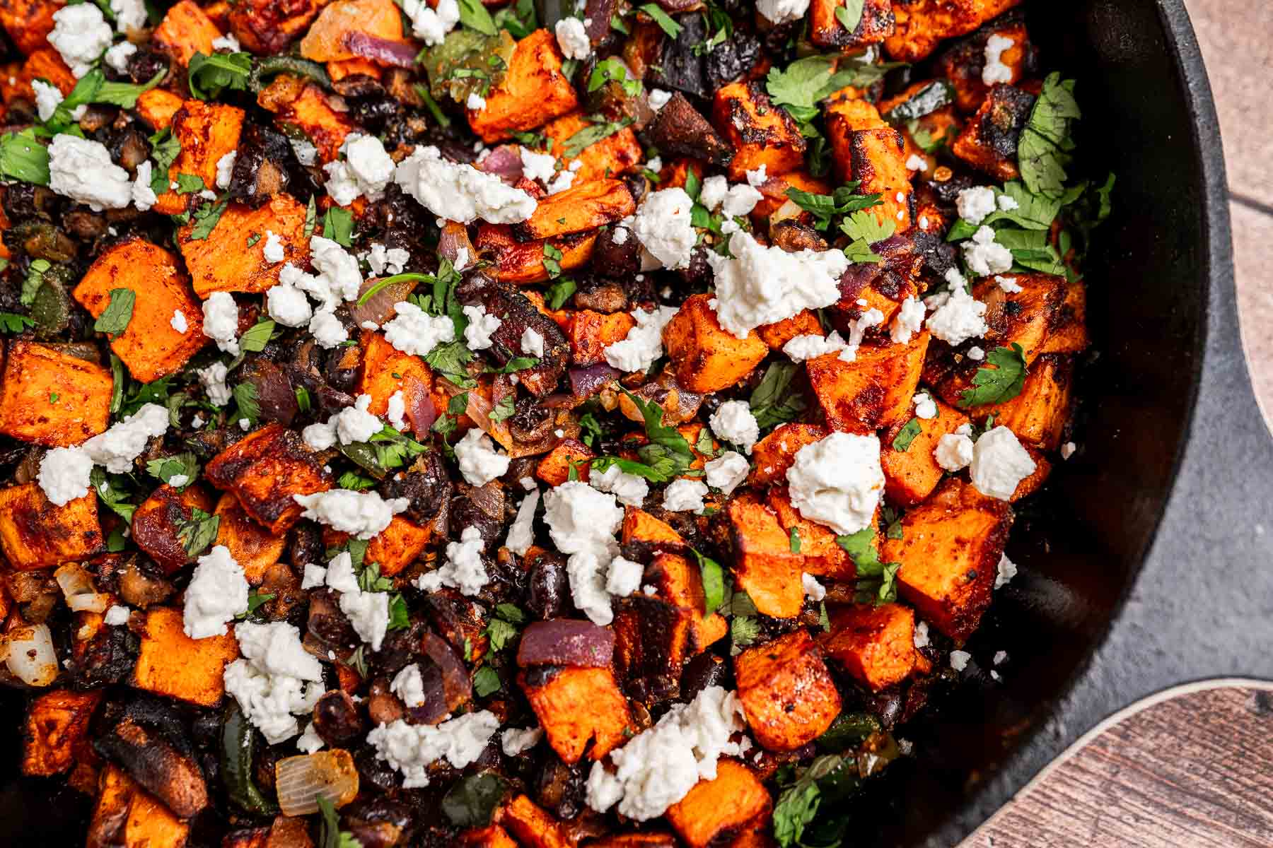 A close-up of a skillet filled with sweet potato hash, featuring roasted sweet potatoes, black beans, chopped herbs, and crumbled feta cheese.