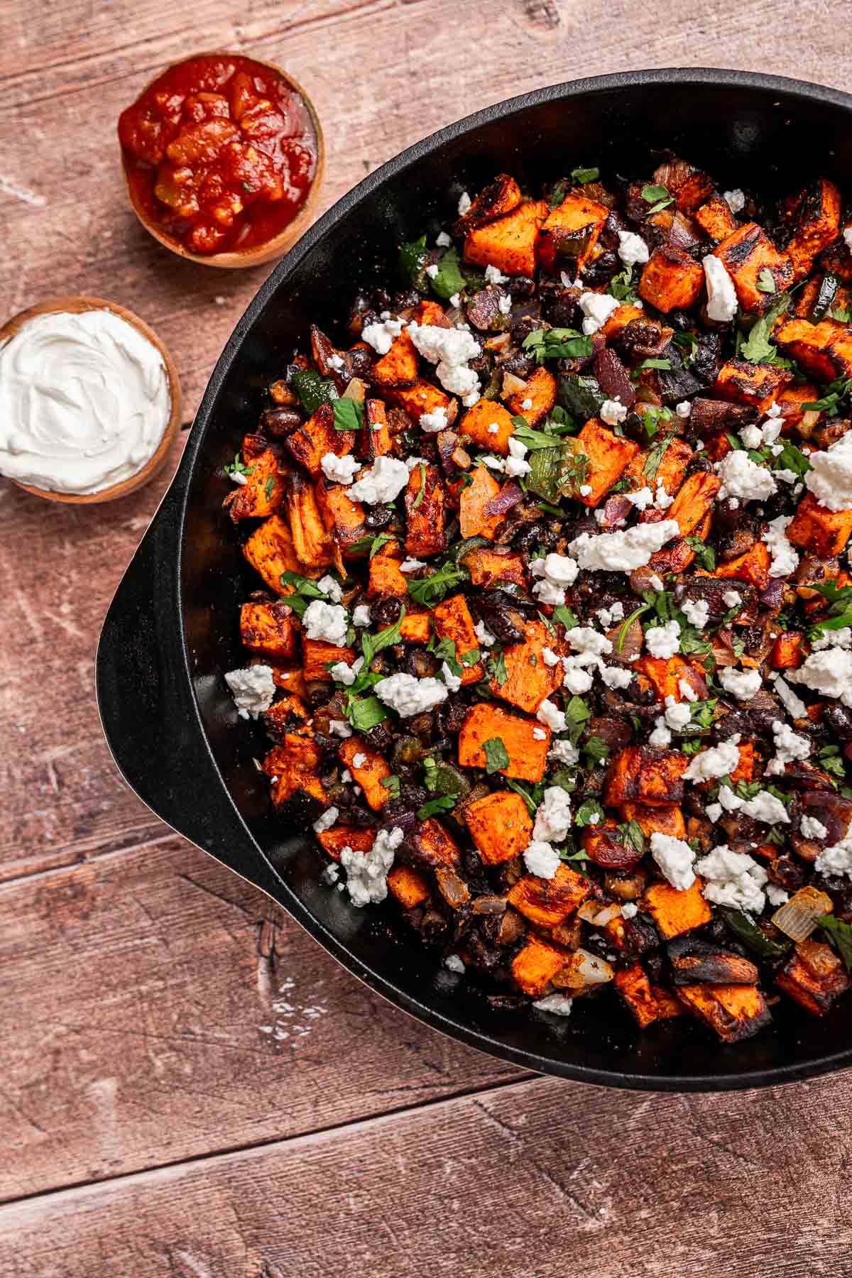 A cast iron skillet filled with sweet potato hash, black beans, herbs, and crumbled cheese. Bowls of salsa and sour cream sit beside the skillet on a wooden surface.