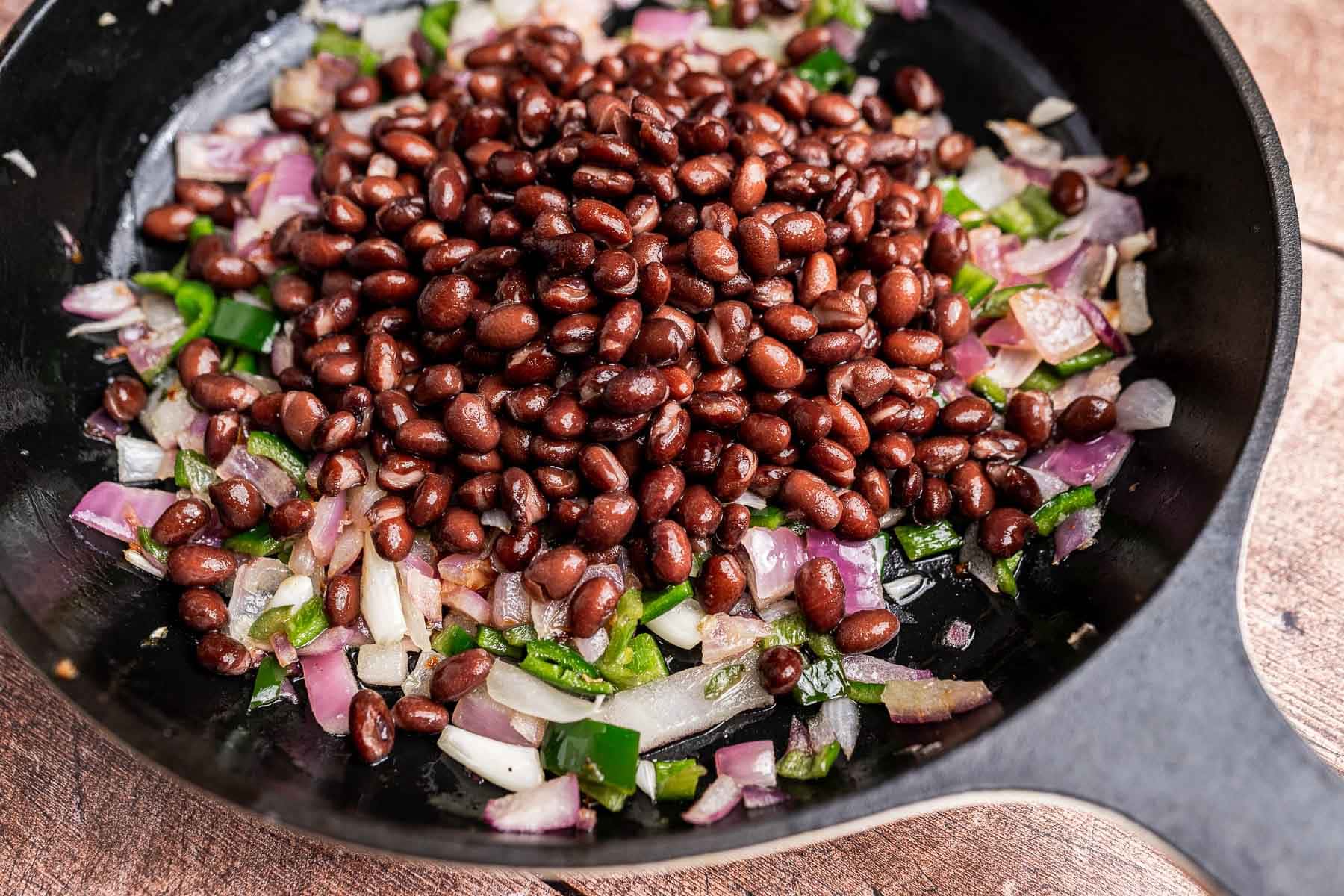 Black beans, chopped onions, and green peppers are being cooked together in a black skillet for a delicious sweet potato hash.